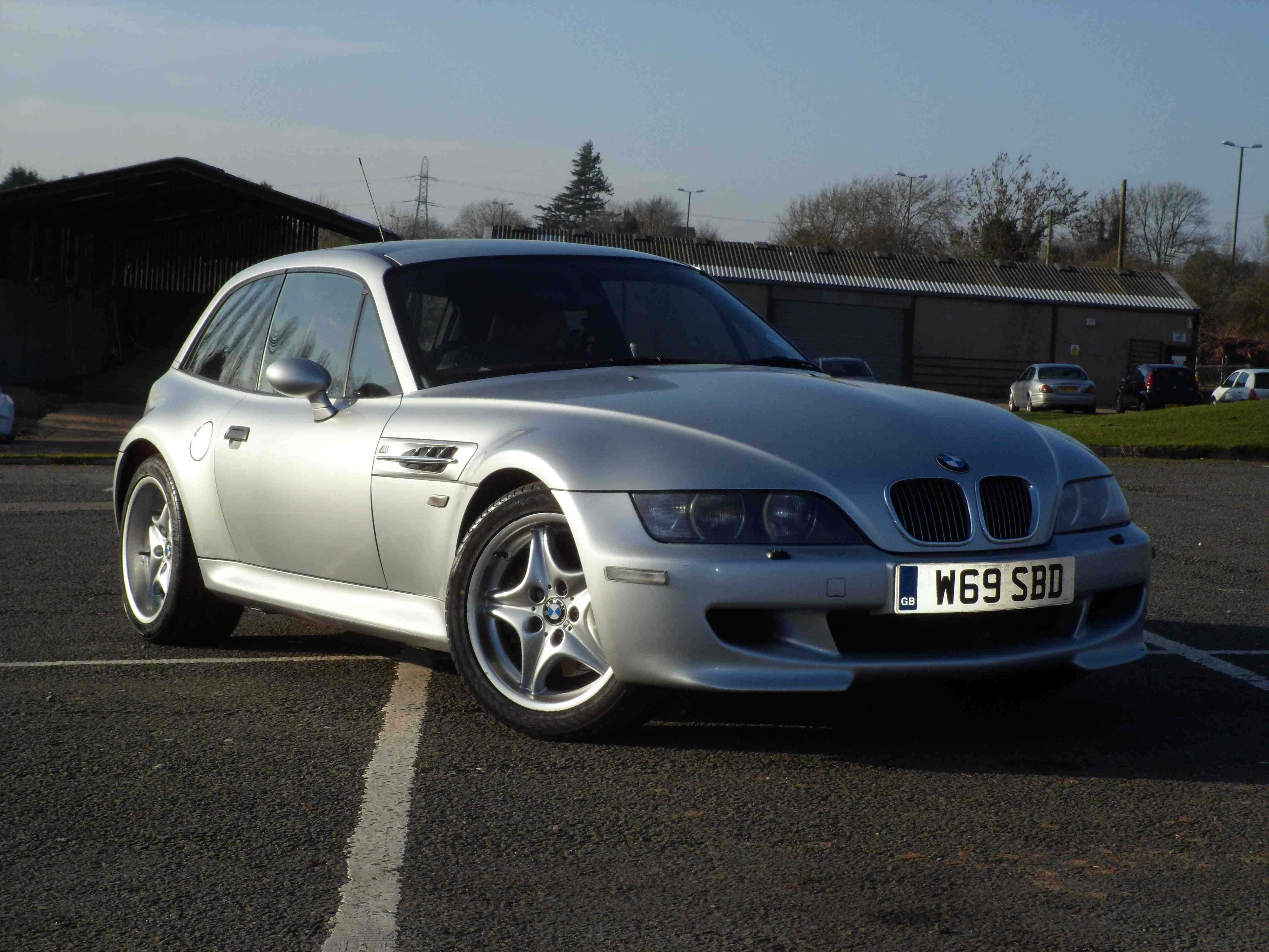 Bmw Z3 桌布 壁紙 — 一台時尚的銀色BMW Z3 M Coupe停放在陽光普照的瀝青停車場，背景有工業建築物和光禿禿的樹木。