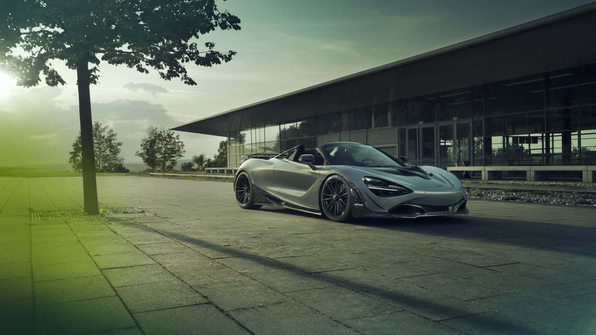 1920 X 1080 Car 桌布 壁紙 — 一台時尚的銀色McLaren Speedtail敞篷跑車停放在鋪砌的廣場上，旁邊是現代玻璃建築，有樹木和柔和的金色陽光。