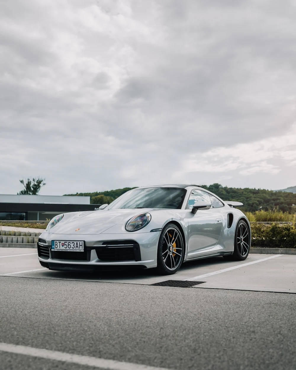 跑車 桌布 壁紙 — 一台時尚的銀色保時捷911 Turbo S停放在空曠的停車場，天空多雲，背景是綠色山丘。
