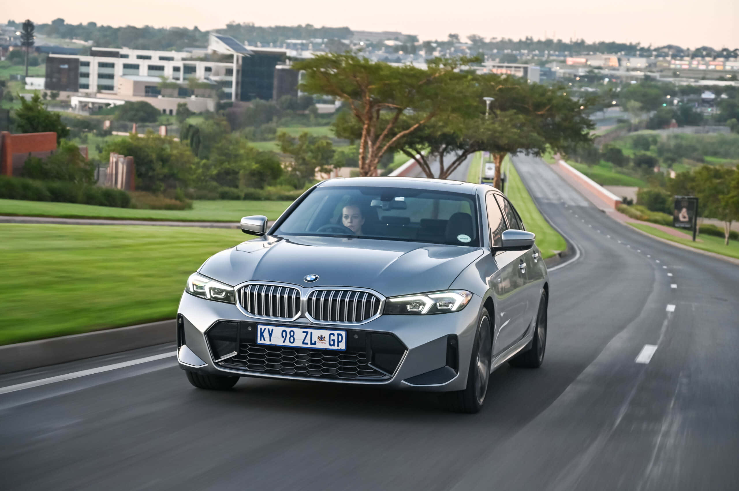 Bmw 3 Series 桌布 壁紙 — 一台時尚的銀色寶馬轎車沿著蜿蜒的道路行駛，穿過柔和的日光下綠色的郊區景觀。