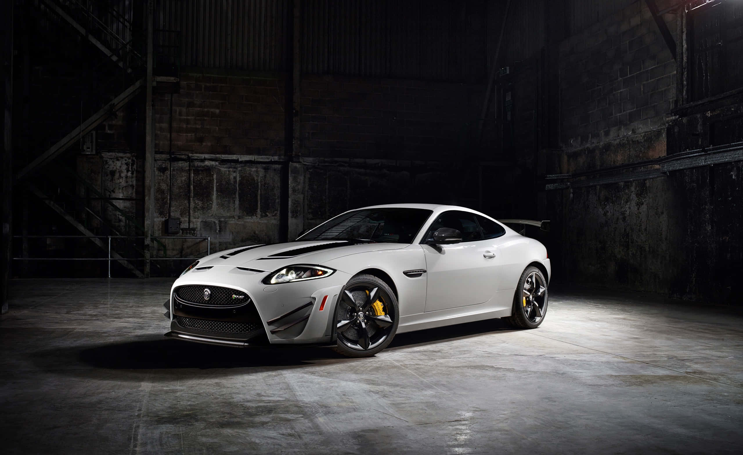Jaguar Xkr 桌布 壁紙 — 一台時尚的銀色捷豹跑車，帶有黑色飾件和黃色制動卡鉗，在骯髒的工業倉庫中被戲劇性地照亮。