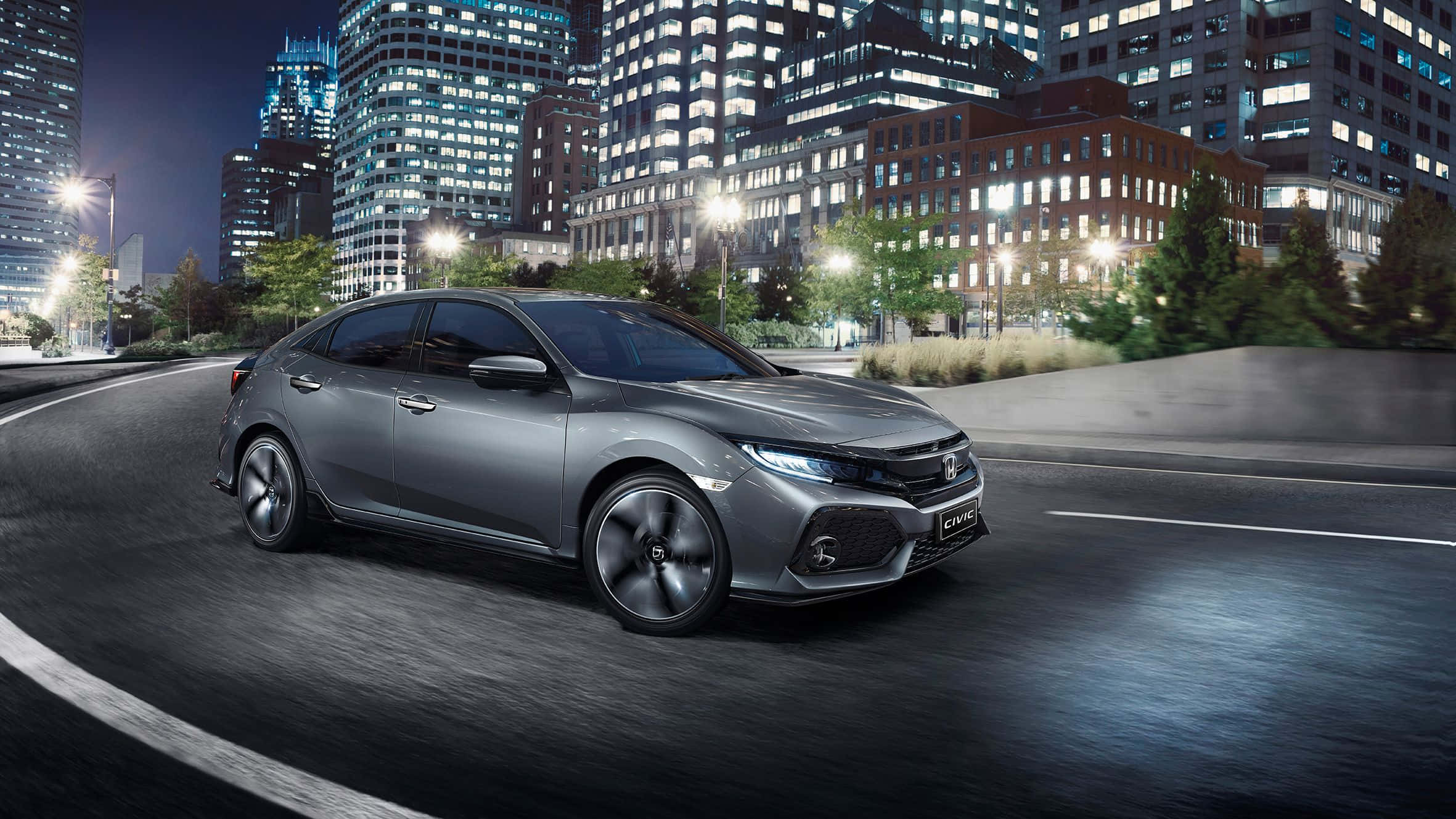 Hatchback 桌布 壁紙 — 一台時尚的銀色本田思域S在夜晚行駛於燈火通明的城市街道，周圍是高聳的摩天大樓。