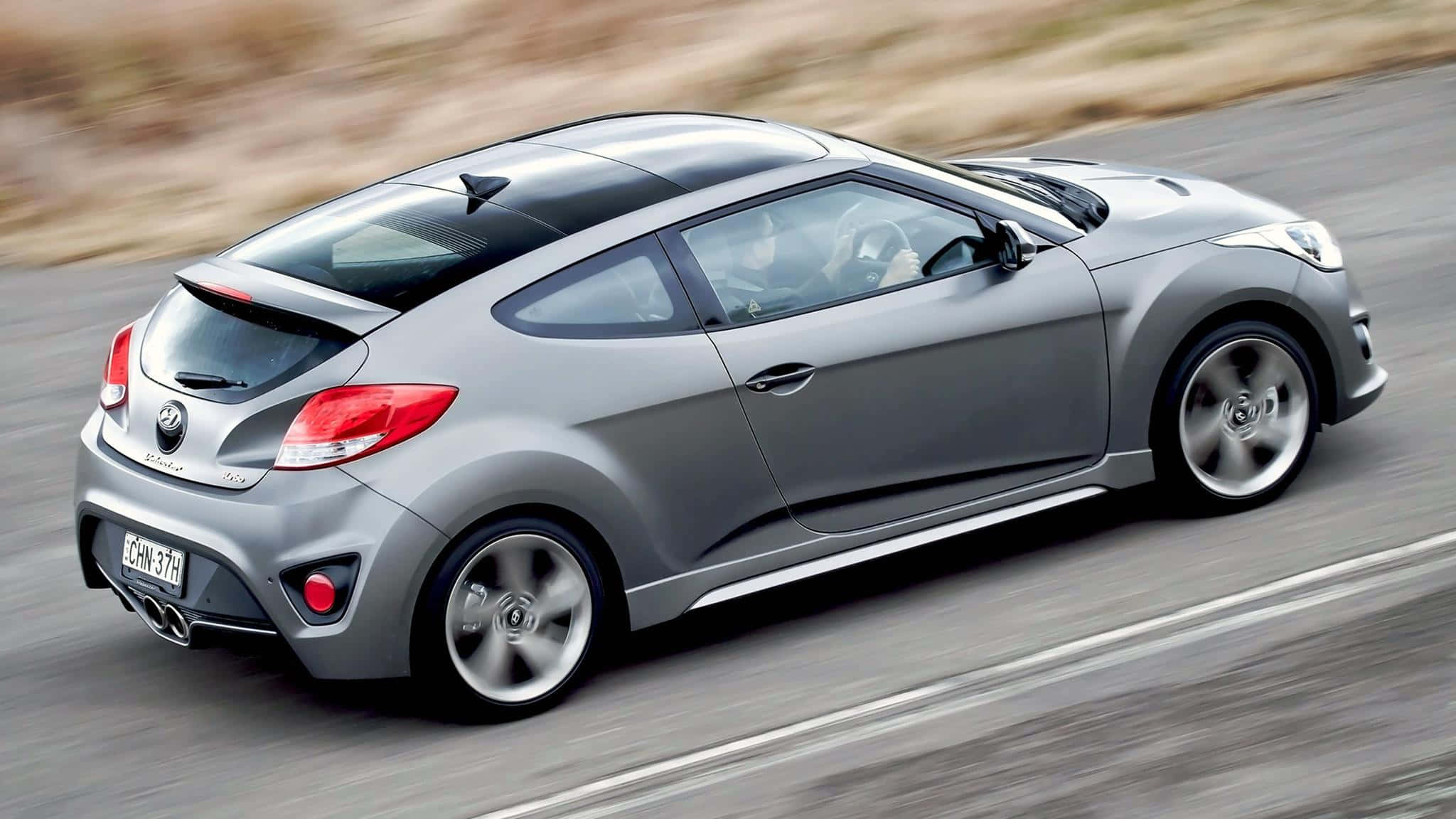 Hyundai Veloster 桌布 壁紙 — 一台時尚的銀色現代Veloster在模糊的道路上疾馳，動態模糊強調了其動感。