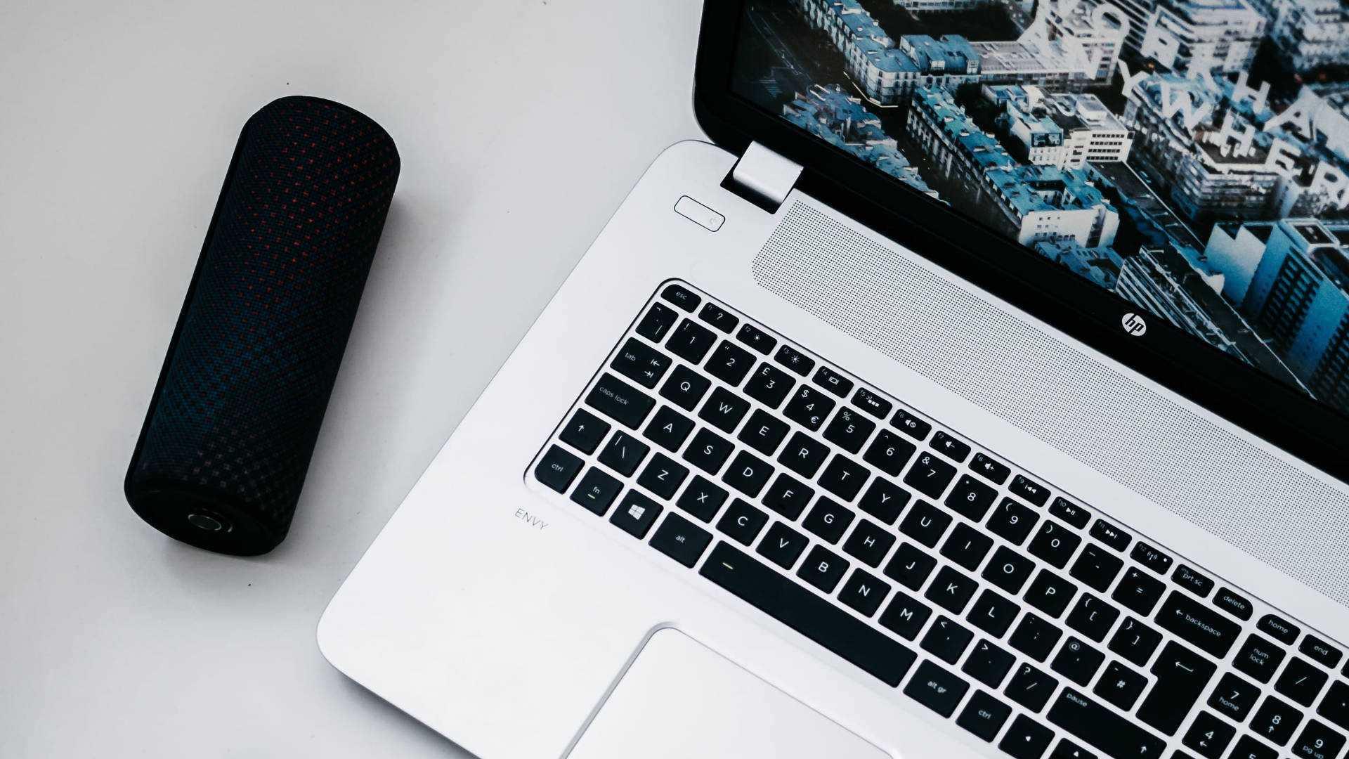 Logitech 桌布 壁紙 — 一台時尚的銀色筆記型電腦，帶有城市景觀桌布，旁邊放著一個黑色圓柱形喇叭，置於簡約的白色表面上。