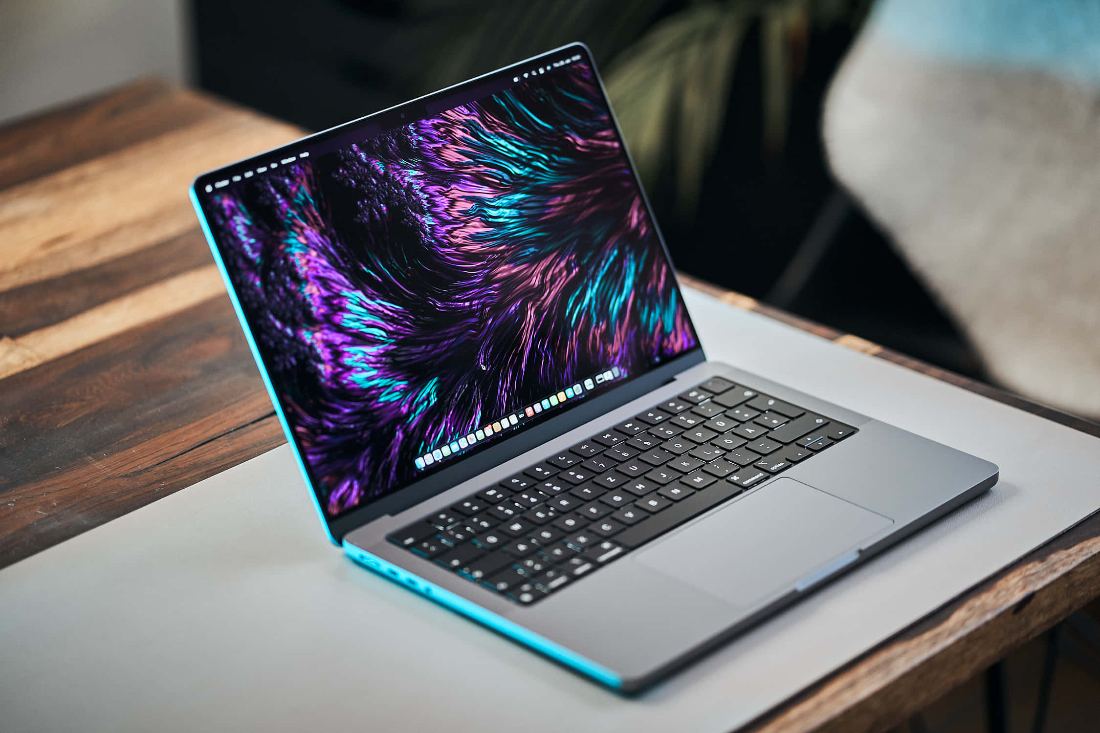 MacBook 桌布 壁紙 — 一台時尚的銀色筆記型電腦，帶有發光的藍色邊緣，在木桌上顯示充滿活力的紫色和藍綠色抽象桌布。