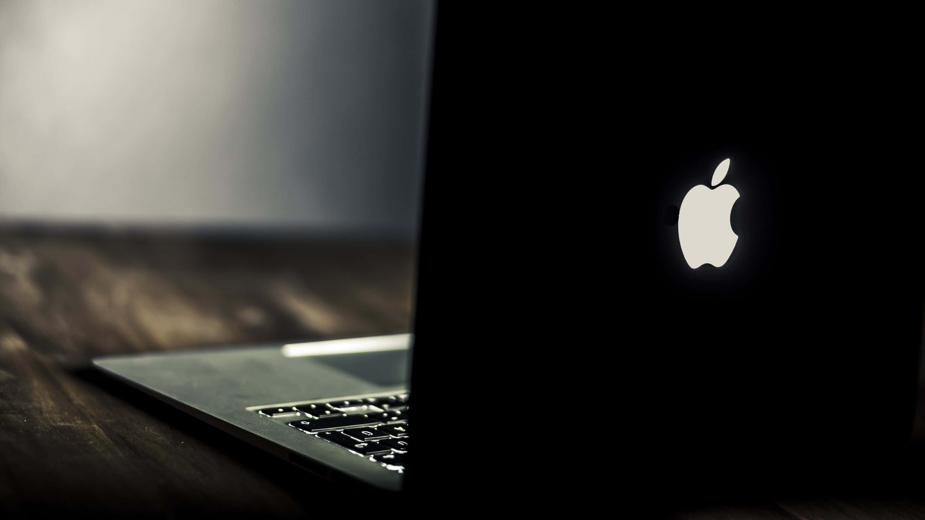 Apple 4K 桌布 壁紙 — 一台時尚的銀色筆記型電腦，帶有發光的Apple標誌，放置在昏暗光線下的深色木質表面上。