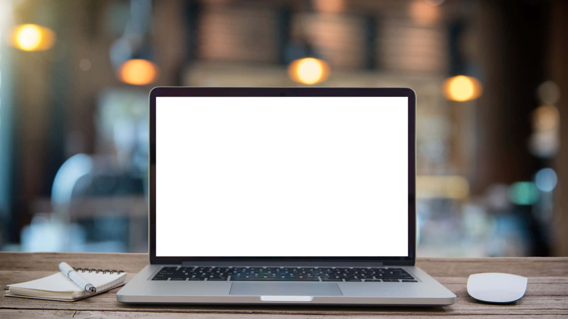 Computer Screen 桌布 壁紙 — 一台時尚的銀色筆記型電腦，配備空白白色螢幕，放在木桌上，旁邊有筆記本和滑鼠，位於燈光溫暖的咖啡館內。