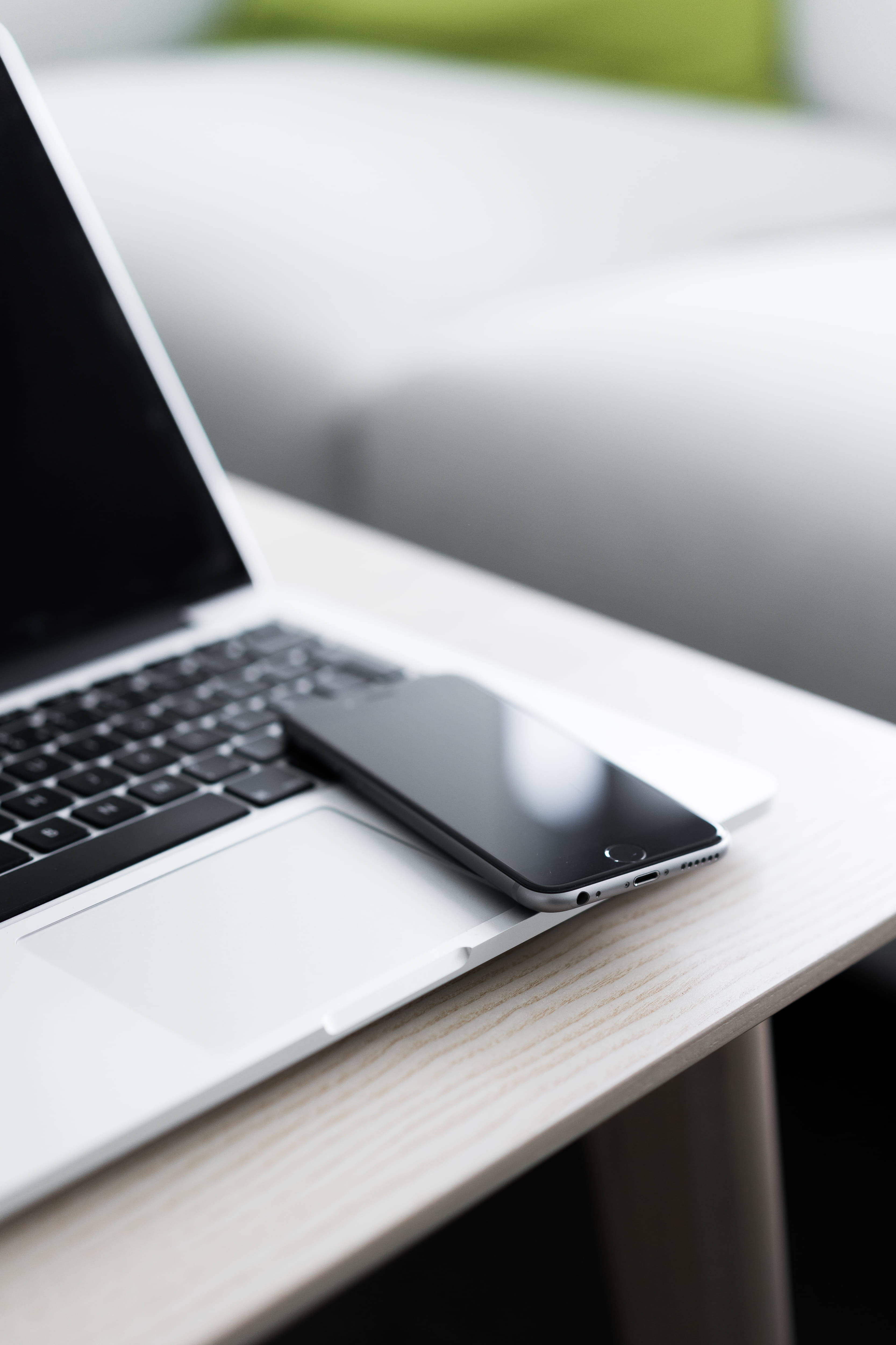 MacBook 動態圖片 桌布 壁紙 — 一台時尚的銀色筆記型電腦和黑色智慧型手機放在淺色木桌上，背景是模糊的白色沙發和綠色枕頭。