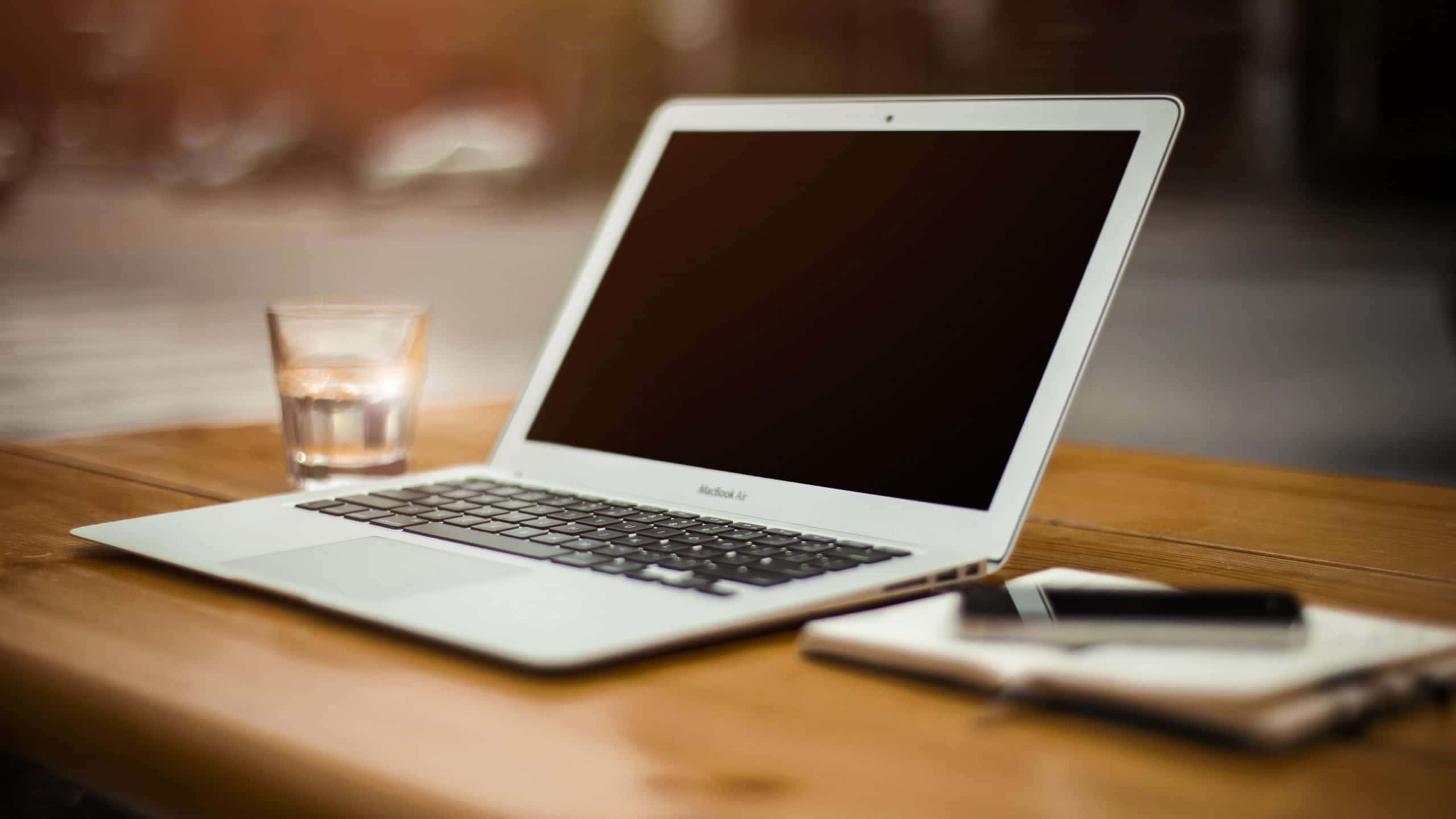 Macbook Air 桌布 壁紙 — 一台時尚的銀色筆記型電腦打開放在溫暖的木桌上，旁邊有一杯水和一本帶筆的筆記本。