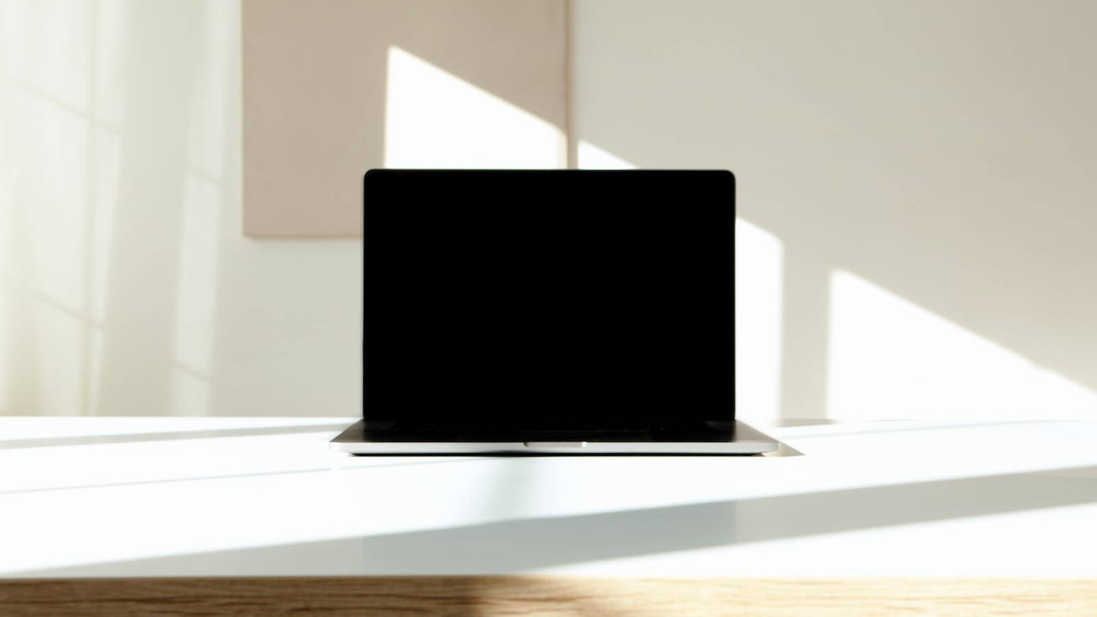 4K Computer 桌布 壁紙 — 一台時尚的銀色筆記型電腦打開放在白色書桌上，沐浴在明亮的自然陽光下，投射出銳利的幾何陰影。
