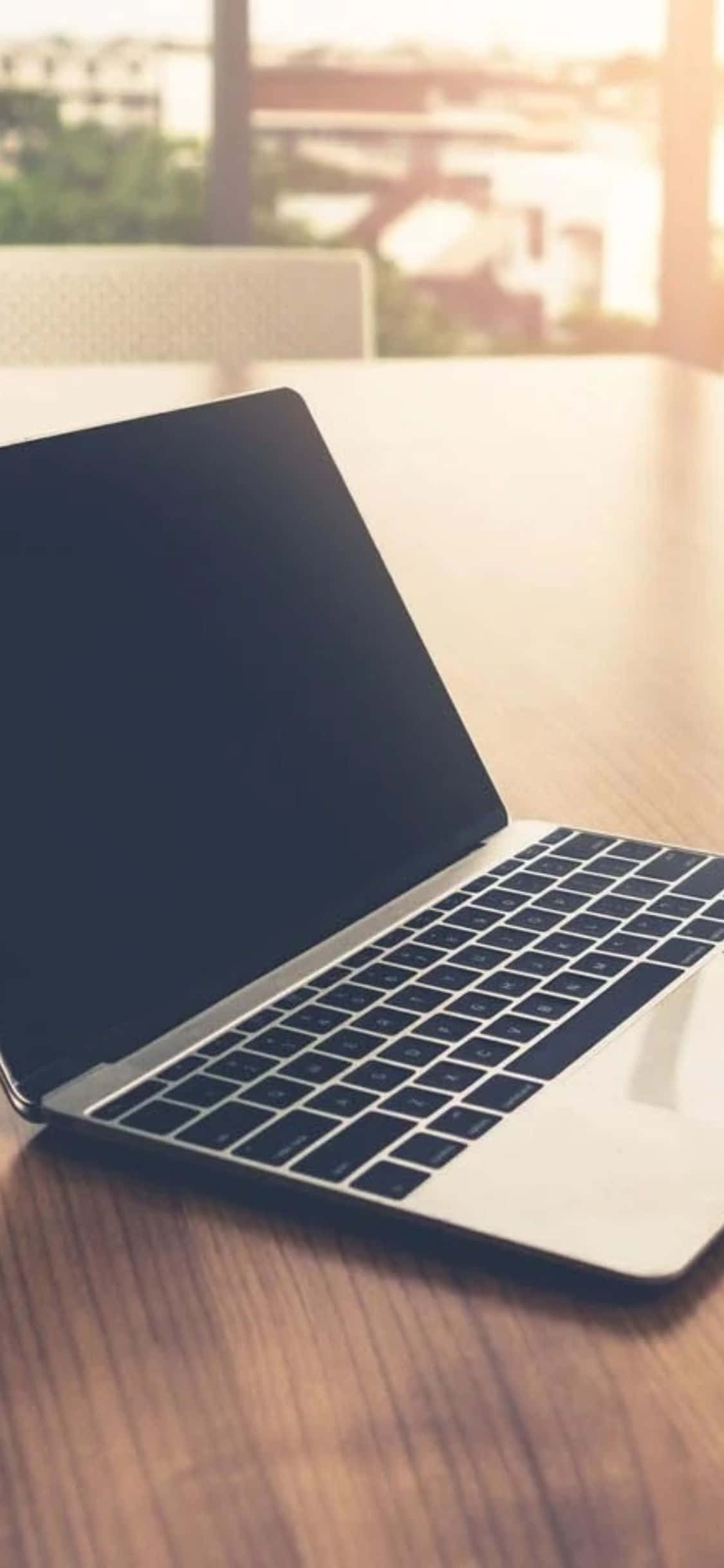 iPhone Xs Computer 桌布 壁紙 — 一台時尚的銀色筆記型電腦打開放在溫暖的木桌上，柔和的金色陽光從窗戶灑進來。