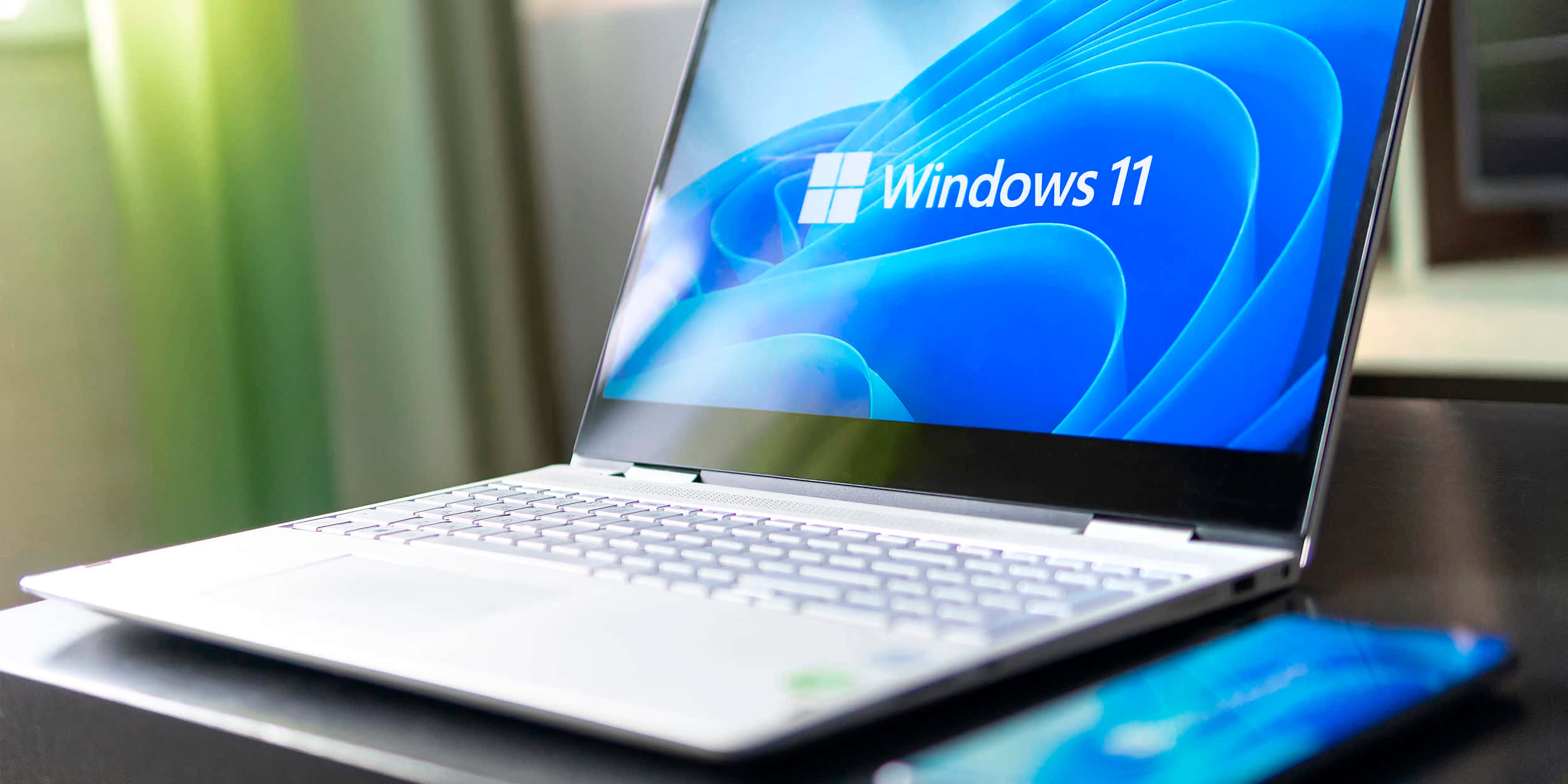 高清 桌布 壁紙 — 一台時尚的銀色筆記型電腦顯示Windows 11啟動畫面，放在深色桌子上，旁邊有一部相配的智慧型手機。