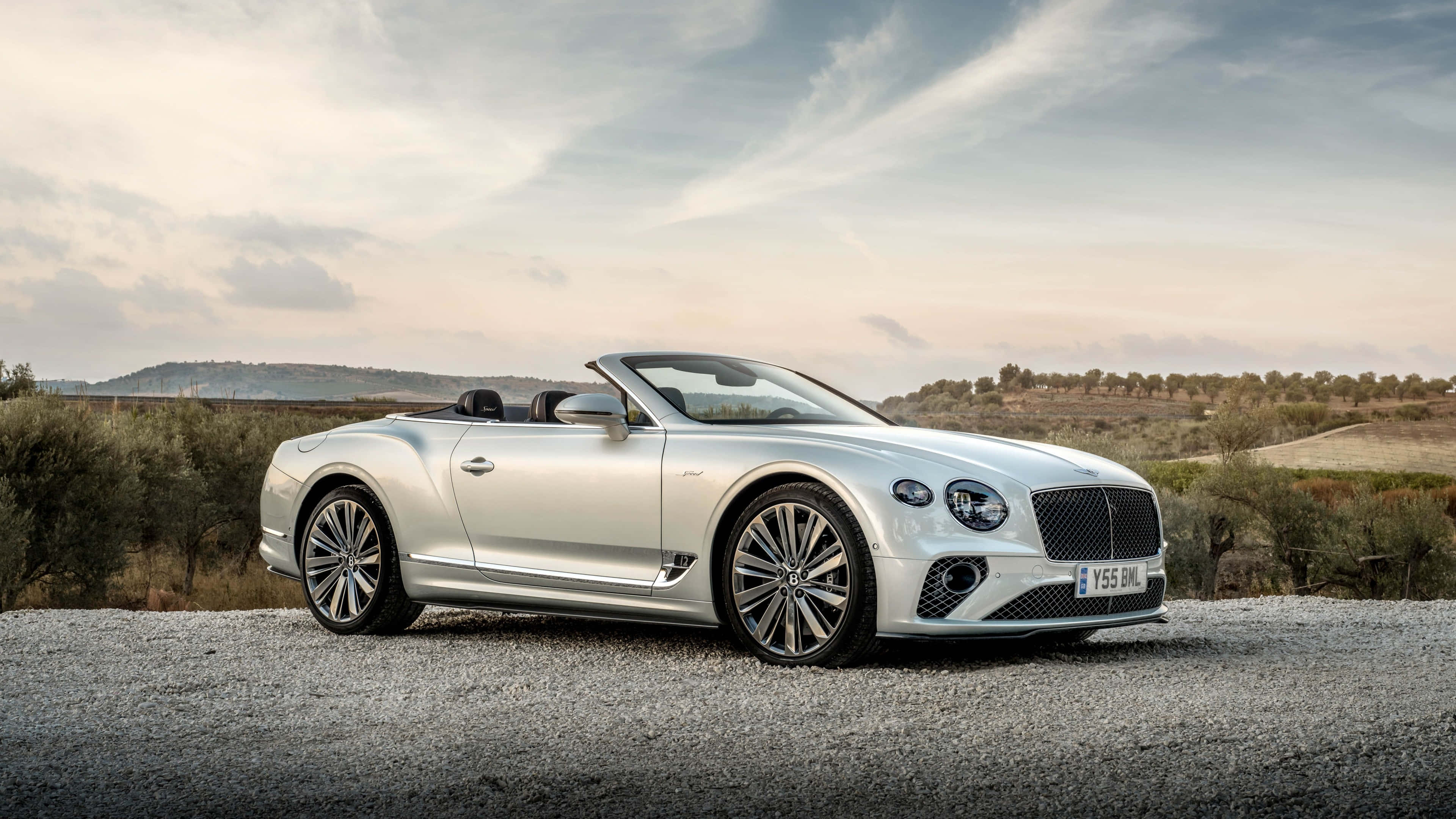 Bentley Continental Gt Convertible 桌布 壁紙 — 一台時尚的銀色賓利敞篷車停在碎石上，背景有起伏的山丘和柔和的雲朵。