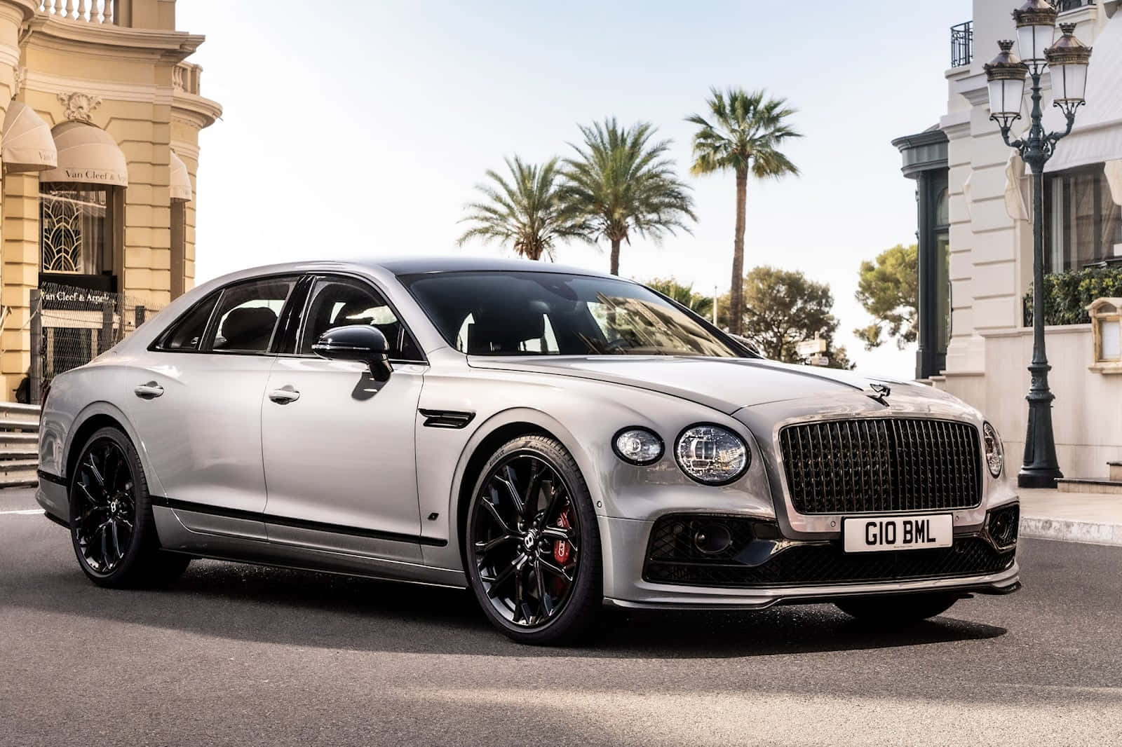 Bentley Flying Spur 桌布 壁紙 — 一台時尚的銀色賓利飛馳，帶有黑色飾邊，停泊在陽光普照的街道上，兩旁種滿棕櫚樹和優雅的建築物。