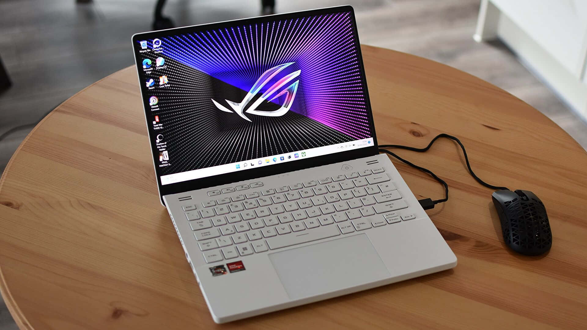 ASUS 桌布 壁紙 — 一台時尚的銀色遊戲筆記型電腦，帶有鮮豔的紫色和黑色 ROG 標誌桌布，打開放在圓形木桌上，旁邊有一個黑色遊戲滑鼠。