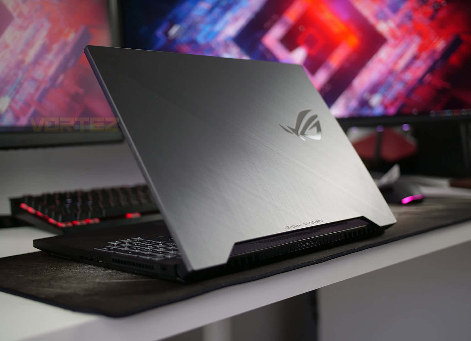 華碩 ROG 桌布 壁紙 — 一台時尚的銀色遊戲筆記型電腦打開放在桌上，背景有發光的紅色鍵盤和鮮豔的RGB顯示器。