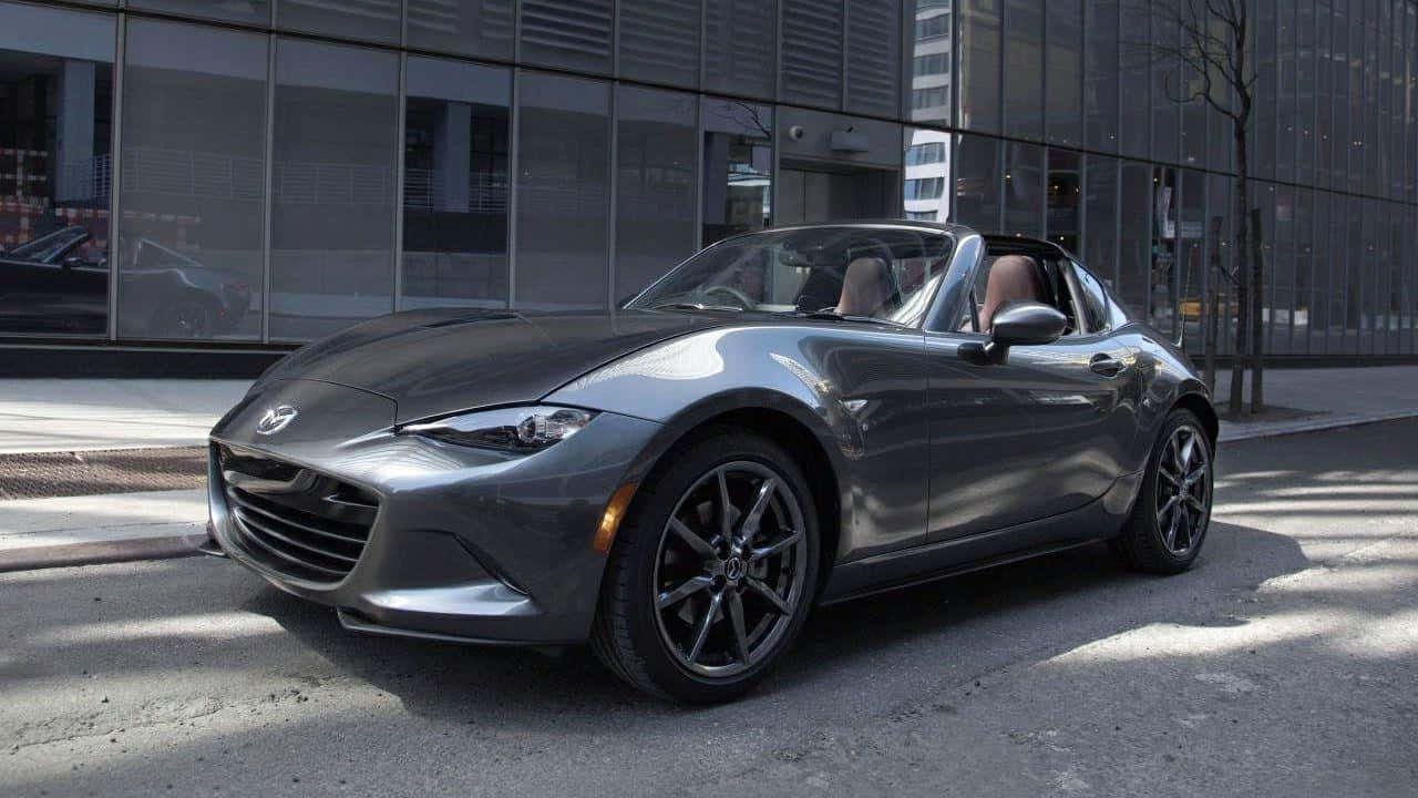 Miata 桌布 壁紙 — 一台時尚的銀色馬自達MX-5 Miata敞篷跑車停放在陽光普照的市街上，旁邊是一座玻璃建築。