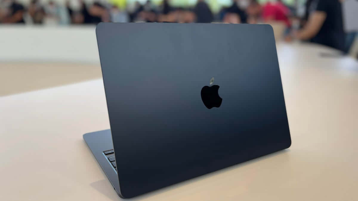 Black Macbook 桌布 壁紙 — 一台時尚的霧面黑色 MacBook 打開放在淺色木桌上，背景有模糊的人群。