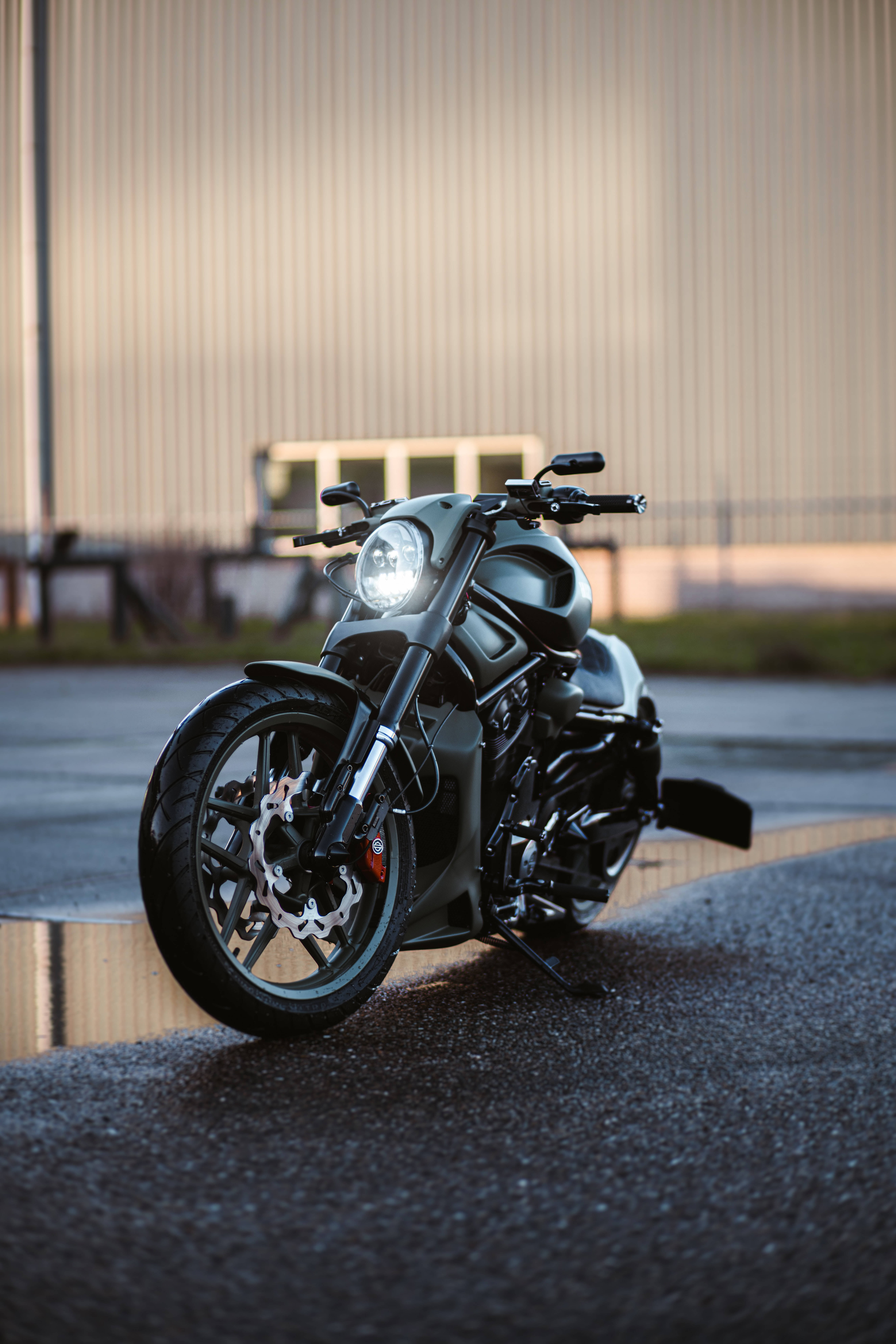 Harley 桌布 壁紙 — 一台時尚的霧面黑色機車，車頭燈發光，停泊在金色時分的瀝青路上，前面是波浪形金屬建築。