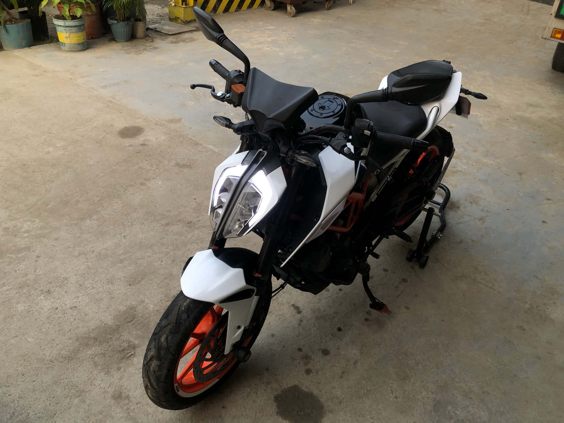 機車 桌布 壁紙 — 一台時尚的黑白機車帶有橙色點綴，停放在車庫的混凝土地板上，附近有盆栽植物。