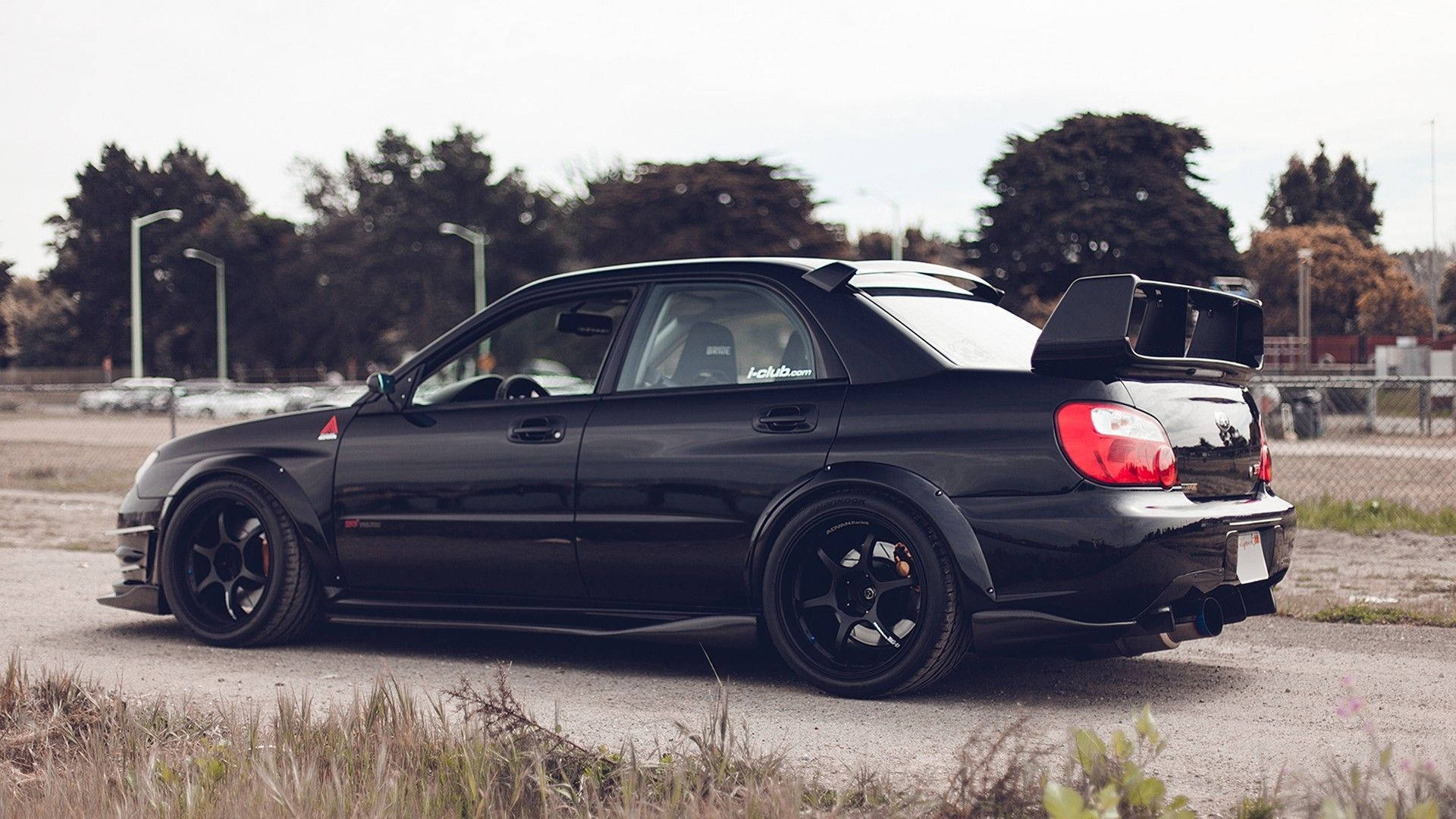 昴 桌布 壁紙 — 一台時尚的黑色 Subaru Impreza WRX STI 配備大型後擾流板，停放在碎石地上，周圍樹木環繞，天空多雲。