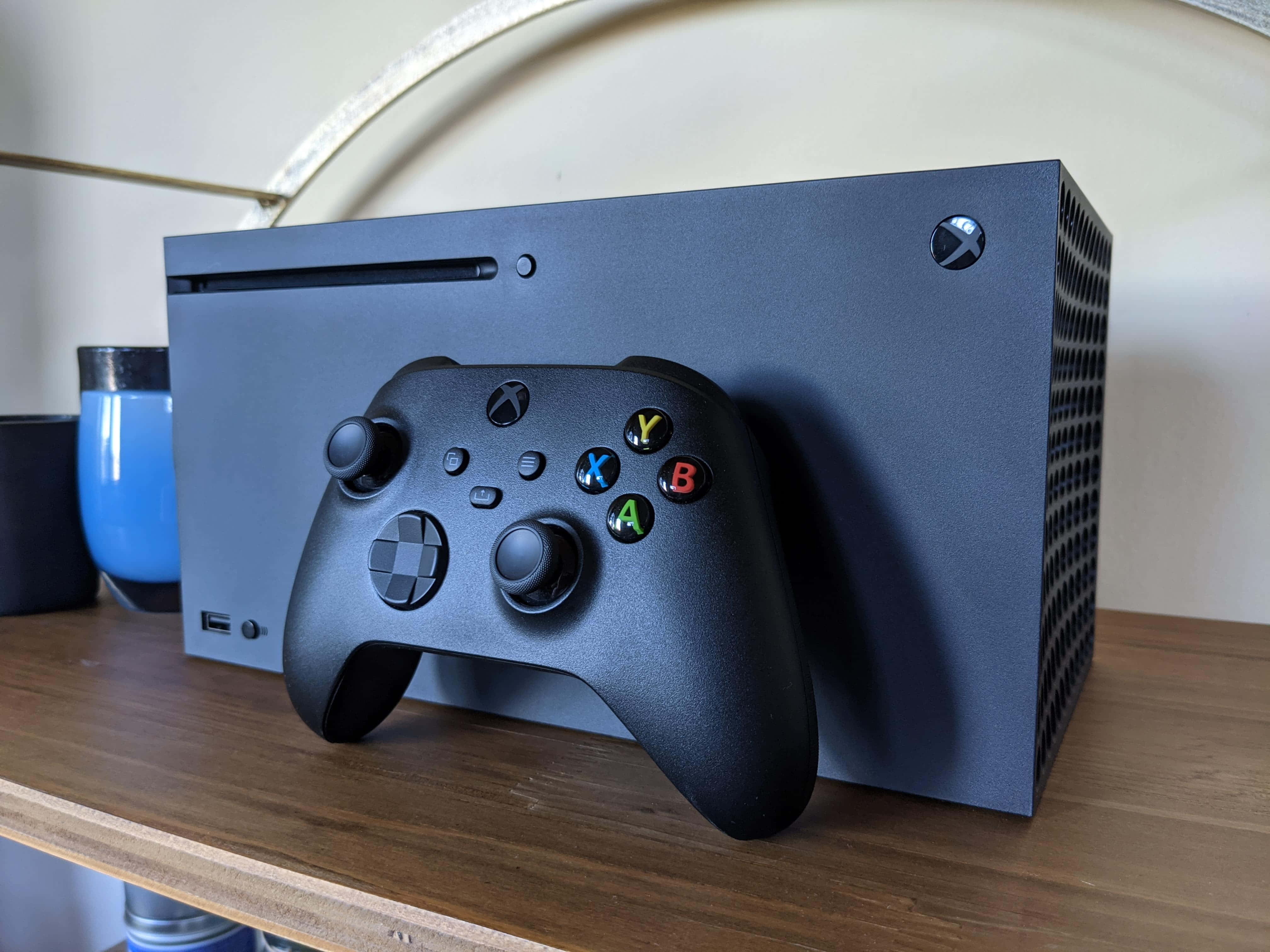 Xbox 桌布 壁紙 — 一台時尚的黑色 Xbox Series X 主機和手把放在木架上，旁邊有一個藍色杯子。