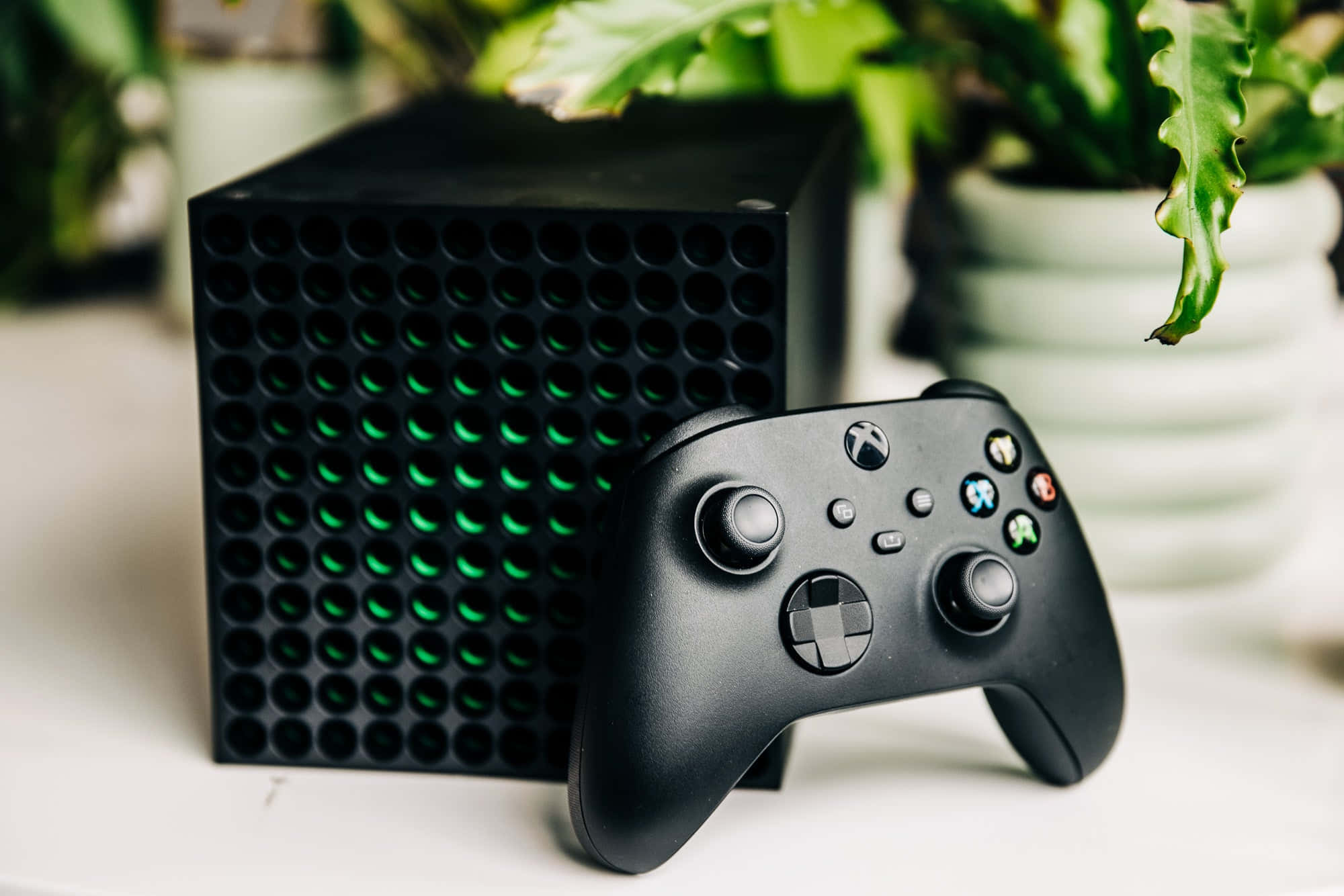 Xbox 桌布 壁紙 — 一台時尚的黑色 Xbox 主機，帶有發光的綠色通風口，與其控制器一起放在白色表面上，周圍環繞著茂盛的綠色植物。