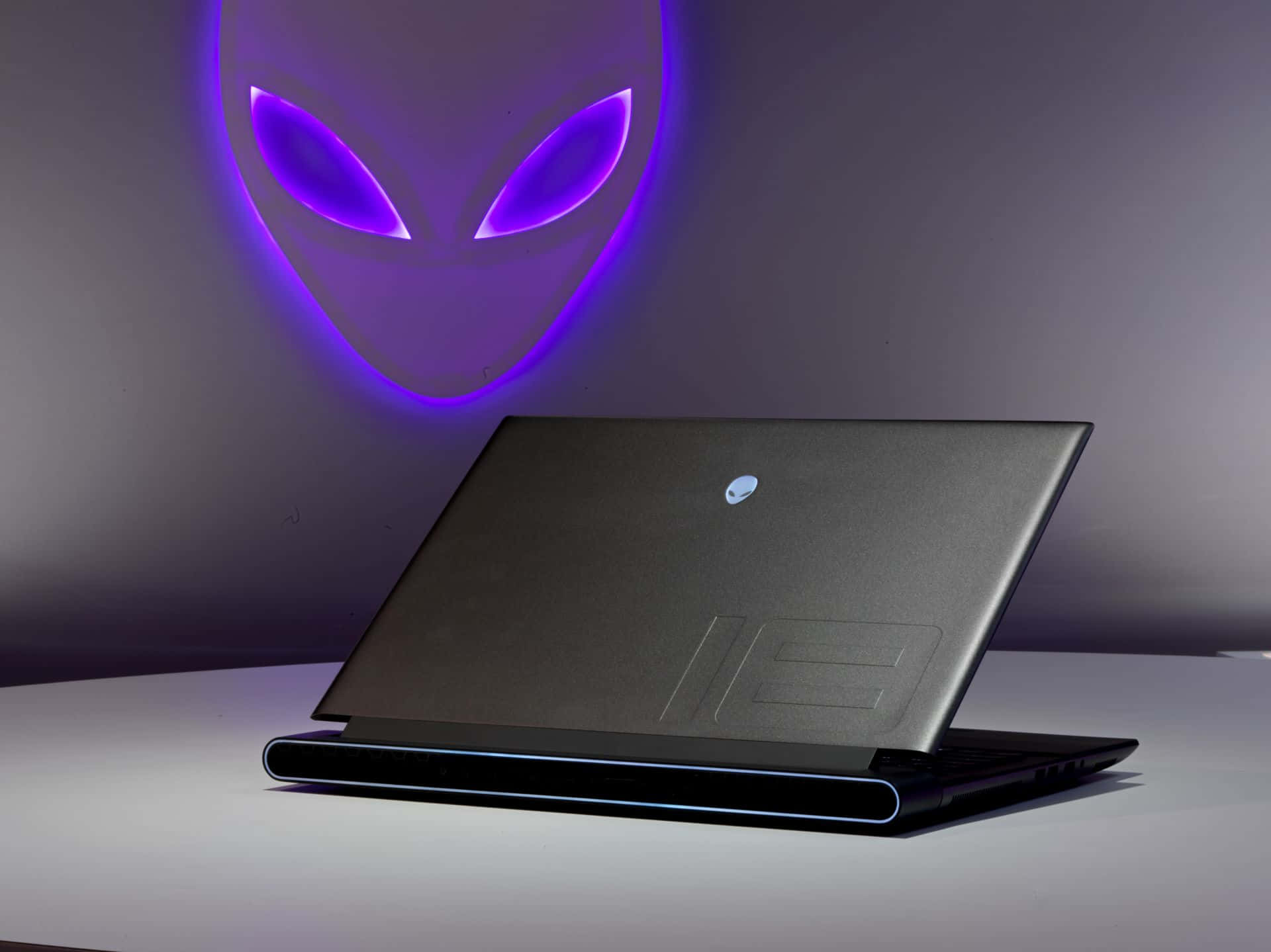 外星人 桌布 壁紙 — 一台時尚的黑色Alienware筆記型電腦放在白色表面上，背後牆上投射出發光的紫色外星人頭標誌。