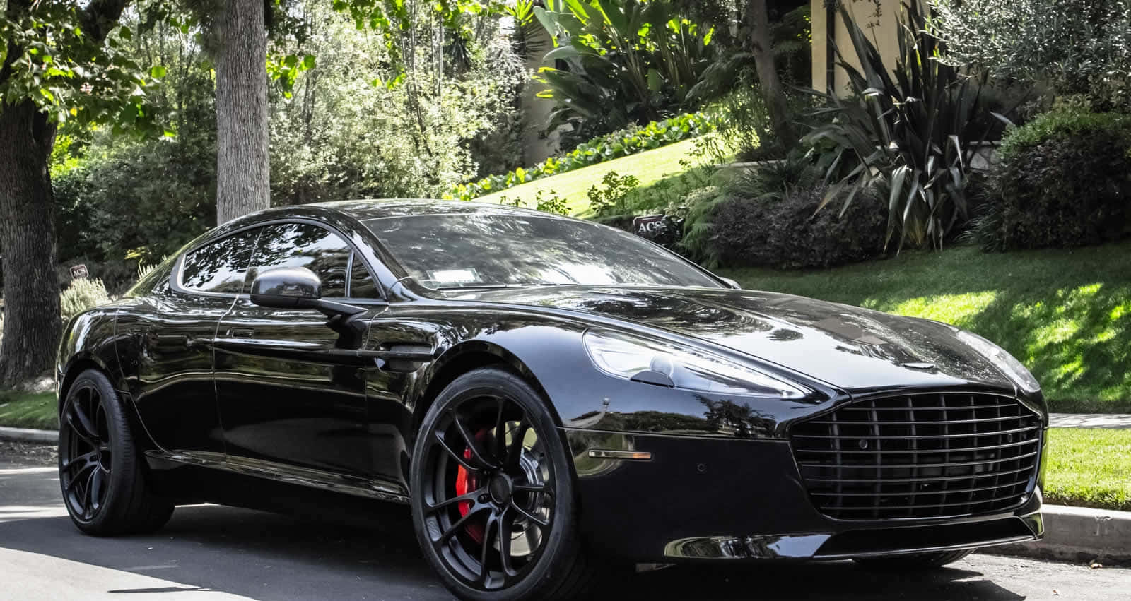 Aston Martin Rapide S 桌布 壁紙 — 一台時尚的黑色Aston Martin跑車在明亮的陽光下閃閃發光，停放在綠樹成蔭的住宅街道上。