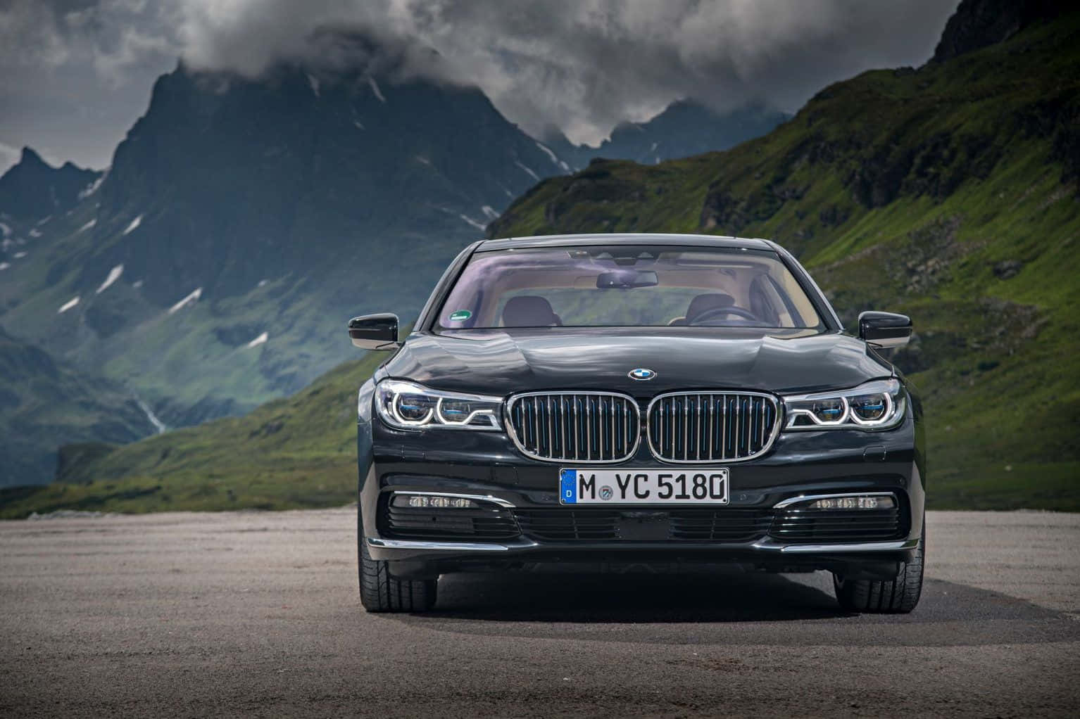 Bmw 7 Series 桌布 壁紙 — 一台時尚的黑色BMW 7系列轎車在山上道路向前行駛，背後有戲劇性的綠色山峰和暴風雲。