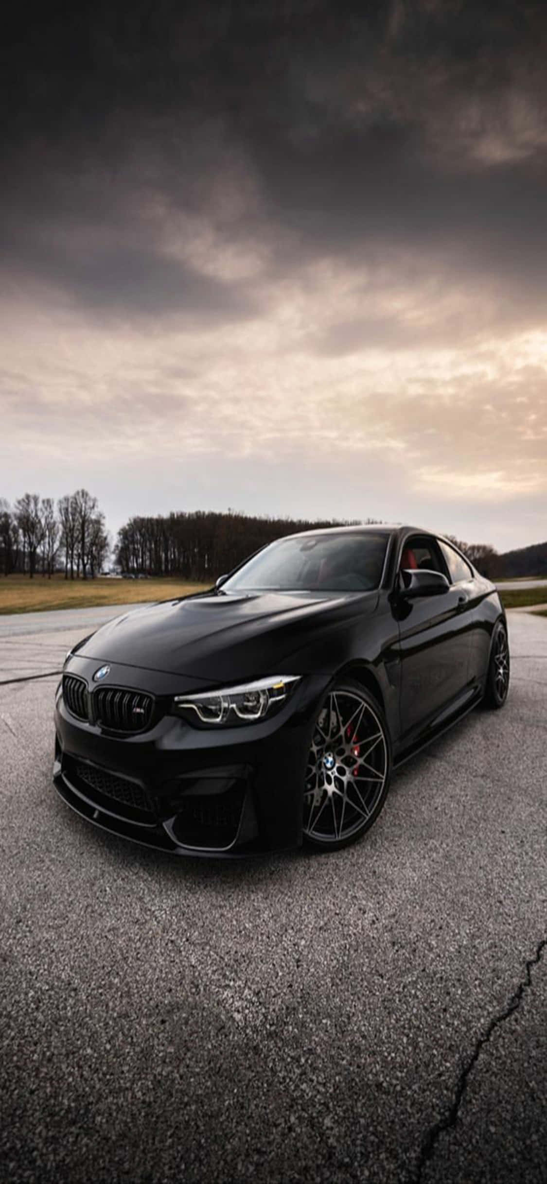 iPhone X Bmw 桌布 壁紙 — 一台時尚的黑色BMW M2雙門跑車停在裂開的柏油路上，背景是黃昏時分戲劇性的多雲天空。