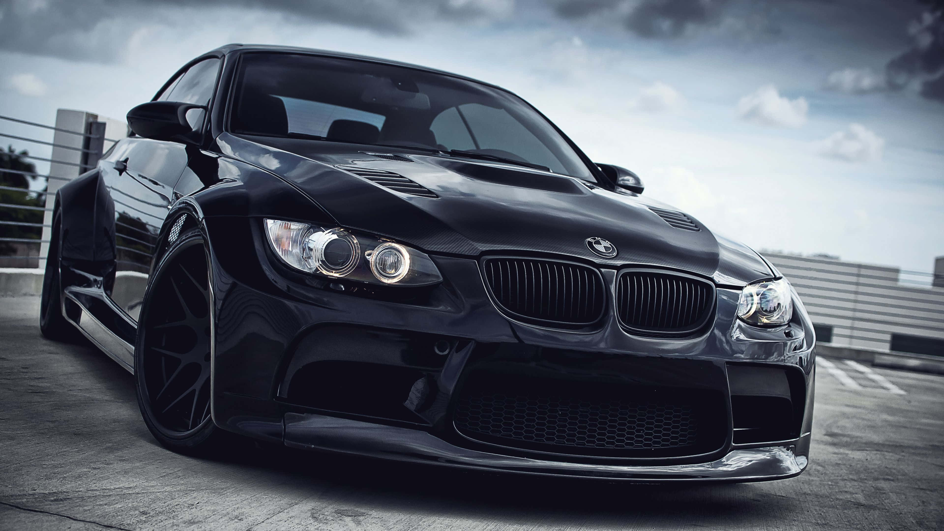 4K BMW 壁紙 桌布 壁紙 — 一台時尚的黑色BMW M3跑車，配備自訂寬體套件和大輪圈，停泊在混凝土屋頂上，天空多雲。