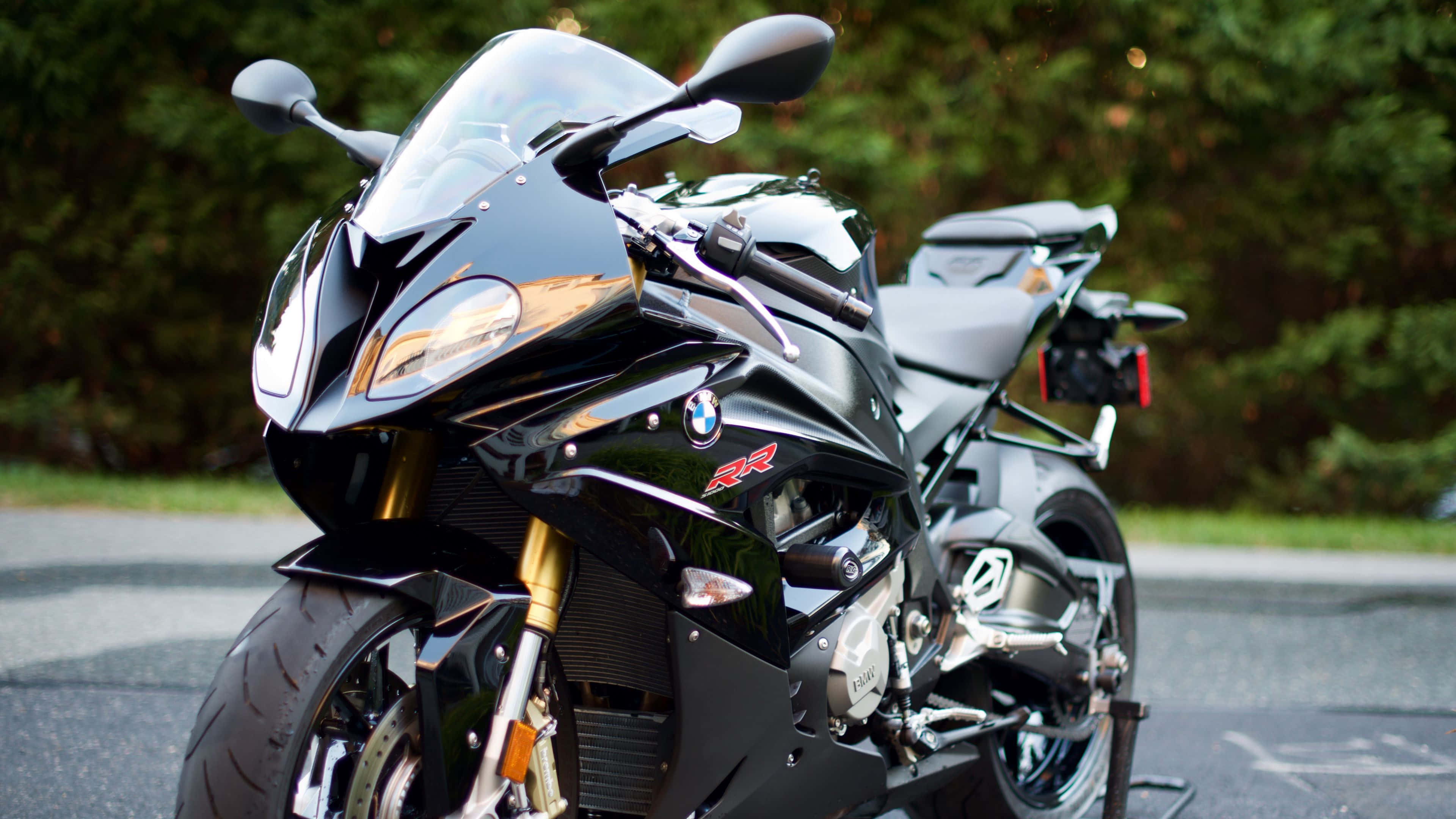 桌布 壁紙 — 一台時尚的黑色BMW R1200RR運動機車在日光下閃耀，配備金色前叉和鍍鉻飾件，停在靠近綠色樹木的柏油路上。