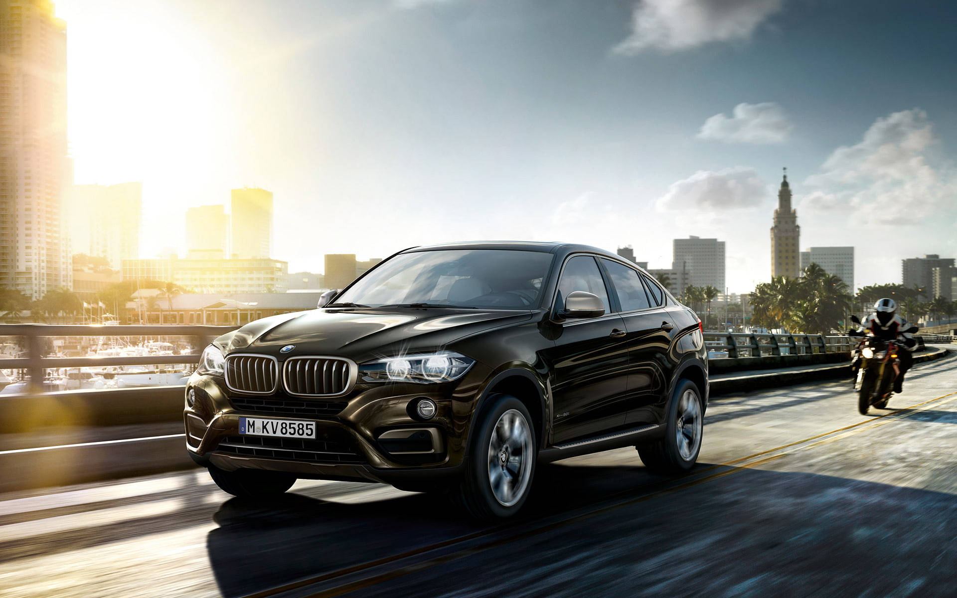 m 桌布 壁紙 — 一台時尚的黑色BMW X6 SUV在陽光明媚的橋上行駛，後面有一位摩托車騎士，背景是城市天際線。