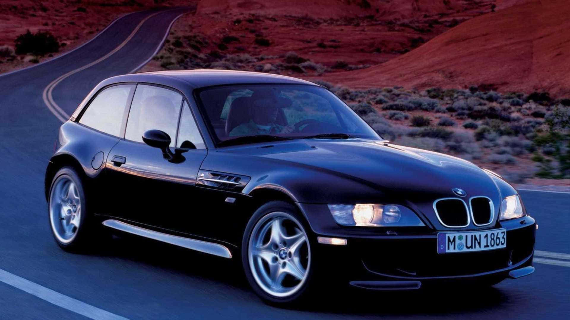 Bmw Z3 桌布 壁紙 — 一台時尚的黑色BMW Z3跑車在日落時分沿著蜿蜒的沙漠公路行駛，背景是紅色岩層。