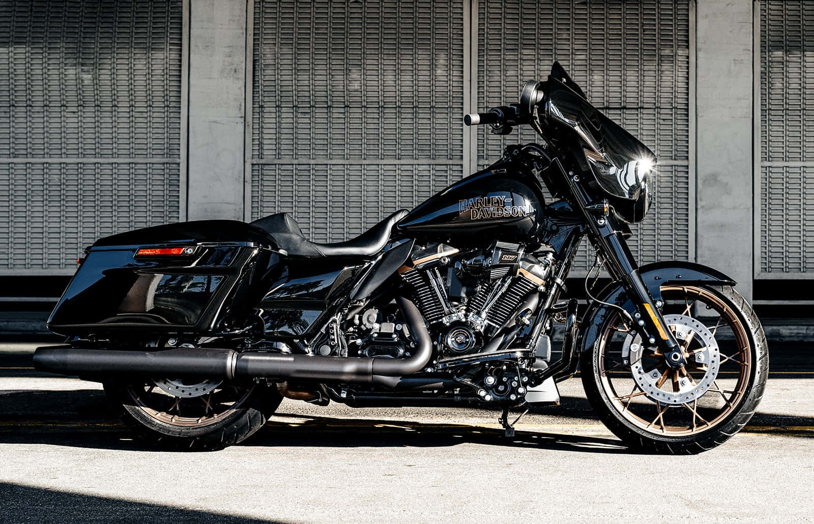哈雷戴維森 桌布 壁紙 — 一台時尚的黑色Harley-Davidson摩托車在明亮陽光下閃閃發光，停放在帶有金屬格柵的混凝土牆前。