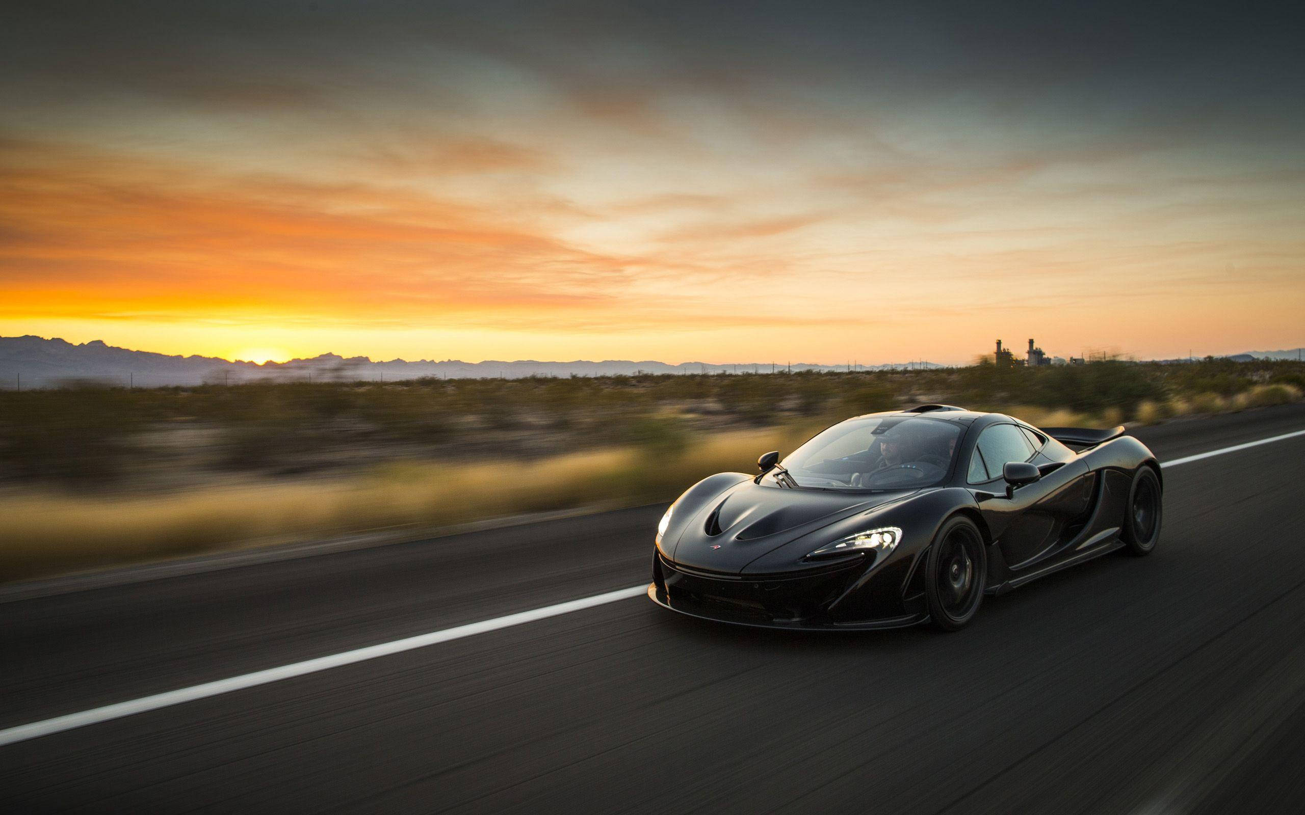 日落 桌布 壁紙 — 一台時尚的黑色McLaren超級跑車在日落時分沿沙漠高速公路疾馳，帶有模糊的動感和溫暖的橙色天空。