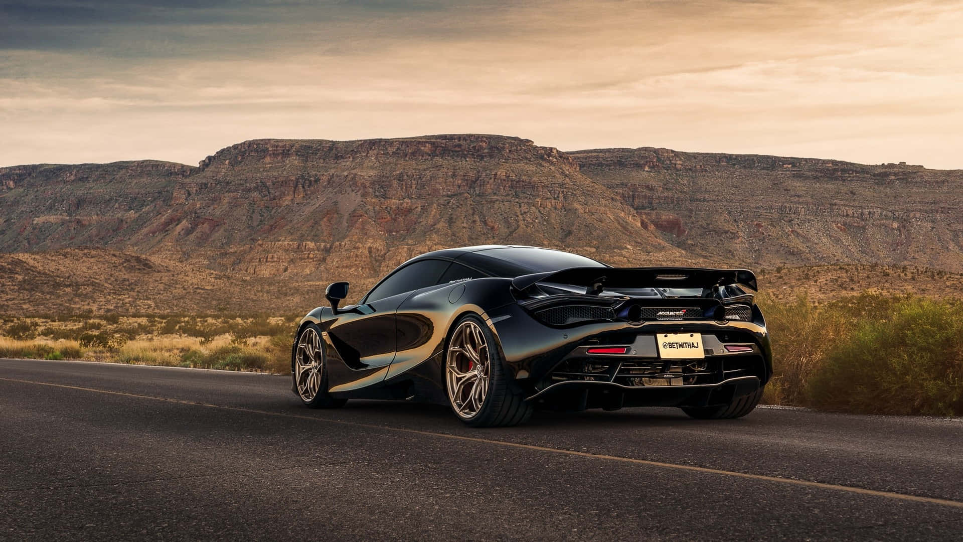 Best Mclaren 720s 桌布 壁紙 — 一台時尚的黑色McLaren跑車沿著沙漠道路行駛，兩旁是崎嶇的懸崖和金色的日落光線。