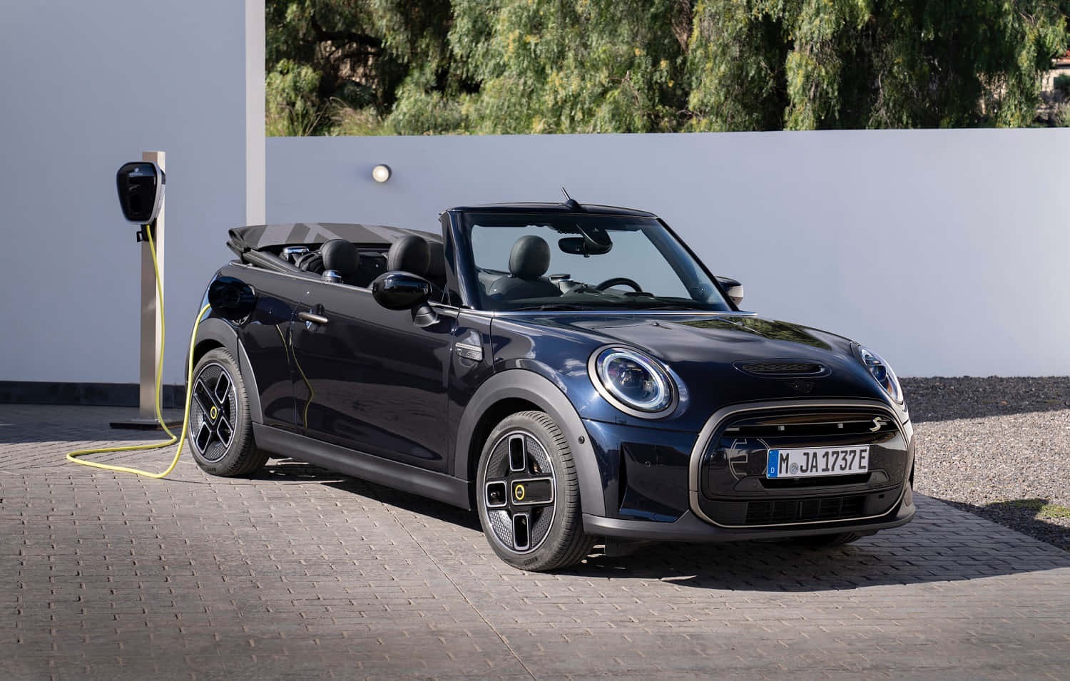 Mini Cooper S Convertible 桌布 壁紙 — 一台時尚的黑色MINI Cooper敞篷車在陽光明媚的日子裡，在白色牆壁後有綠樹的充電站充電。