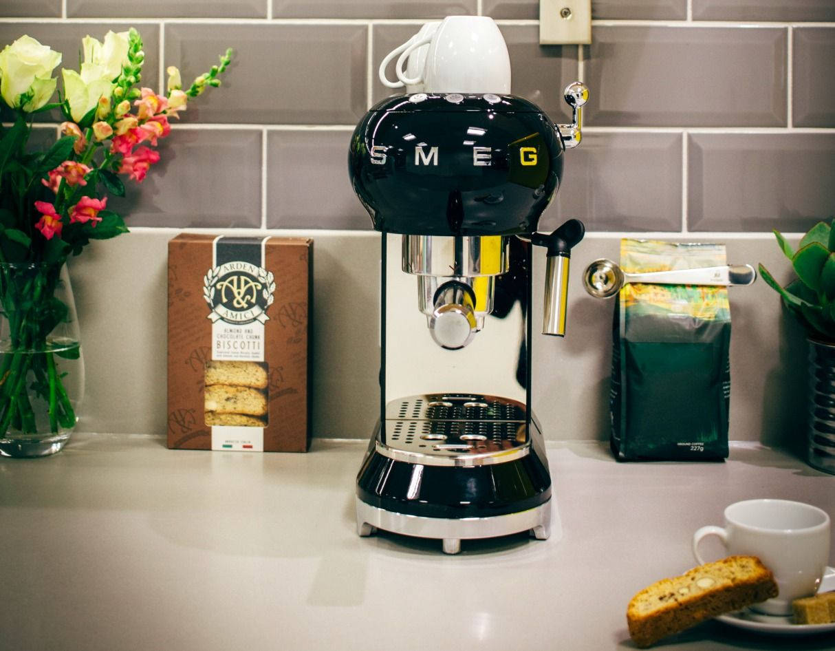 kitchen 桌布 壁紙 — 一台時尚的黑色SMEG濃縮咖啡機放在現代廚房檯面上，旁邊有一瓶色彩繽紛的花和一碟義大利脆餅。