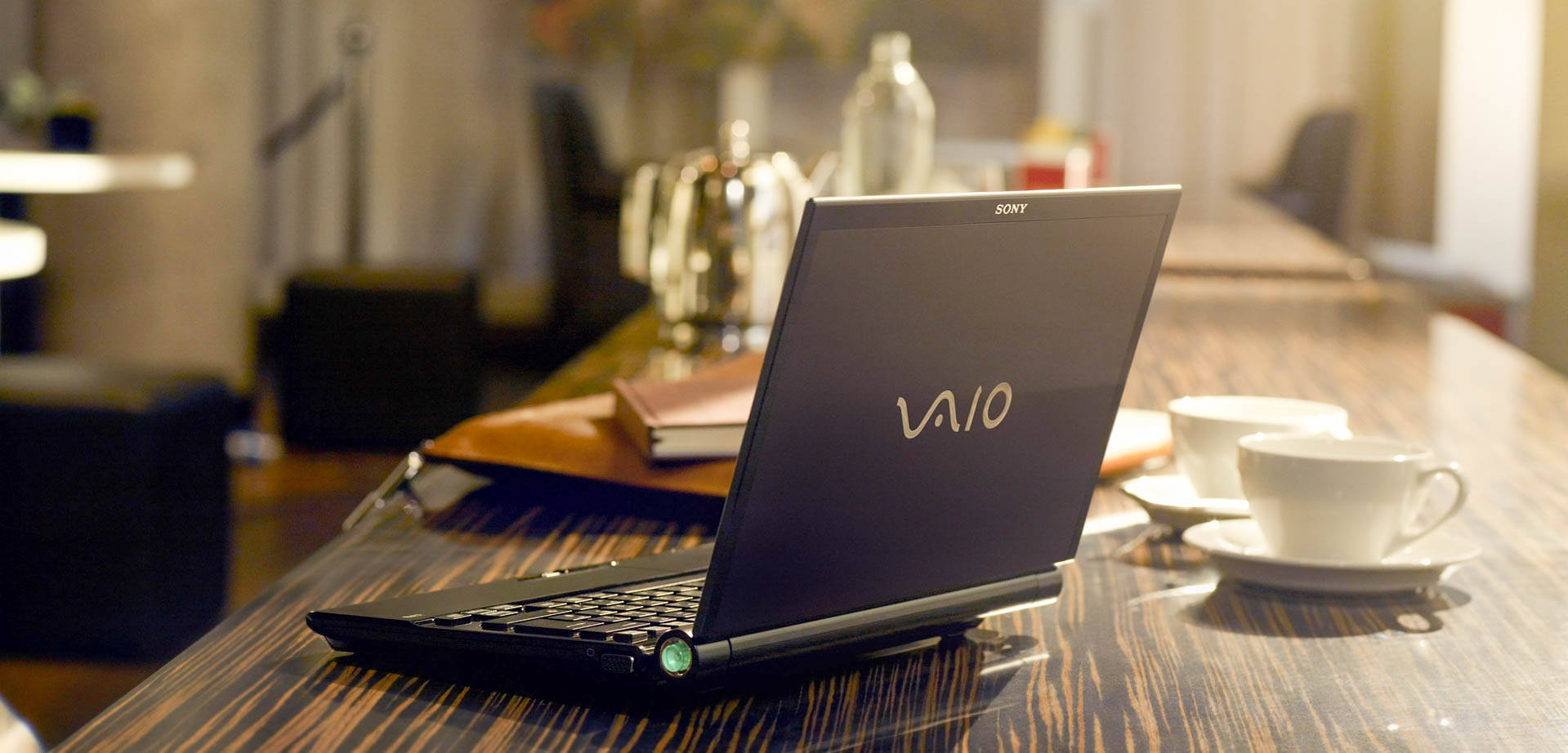 Sony 桌布 壁紙 — 一台時尚的黑色Sony Vaio筆記型電腦打開放在深色木桌上，旁邊有兩個白色咖啡杯，營造出舒適的咖啡館氛圍。