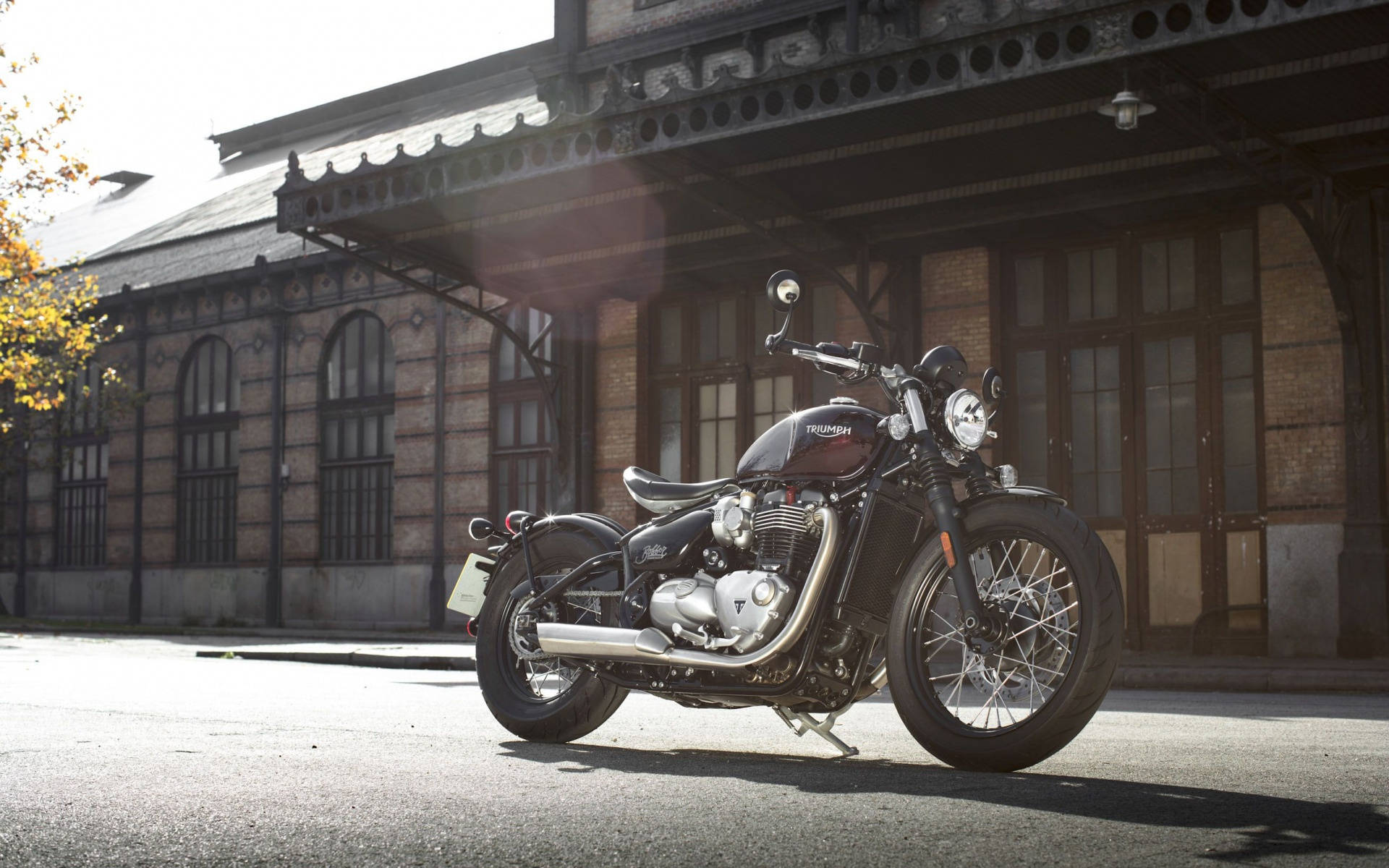 Triumph 桌布 壁紙 — 一台時尚的黑色Triumph摩托車在陽光下閃閃發光，停放在一個帶有華麗鐵藝的歷史磚造火車站前。