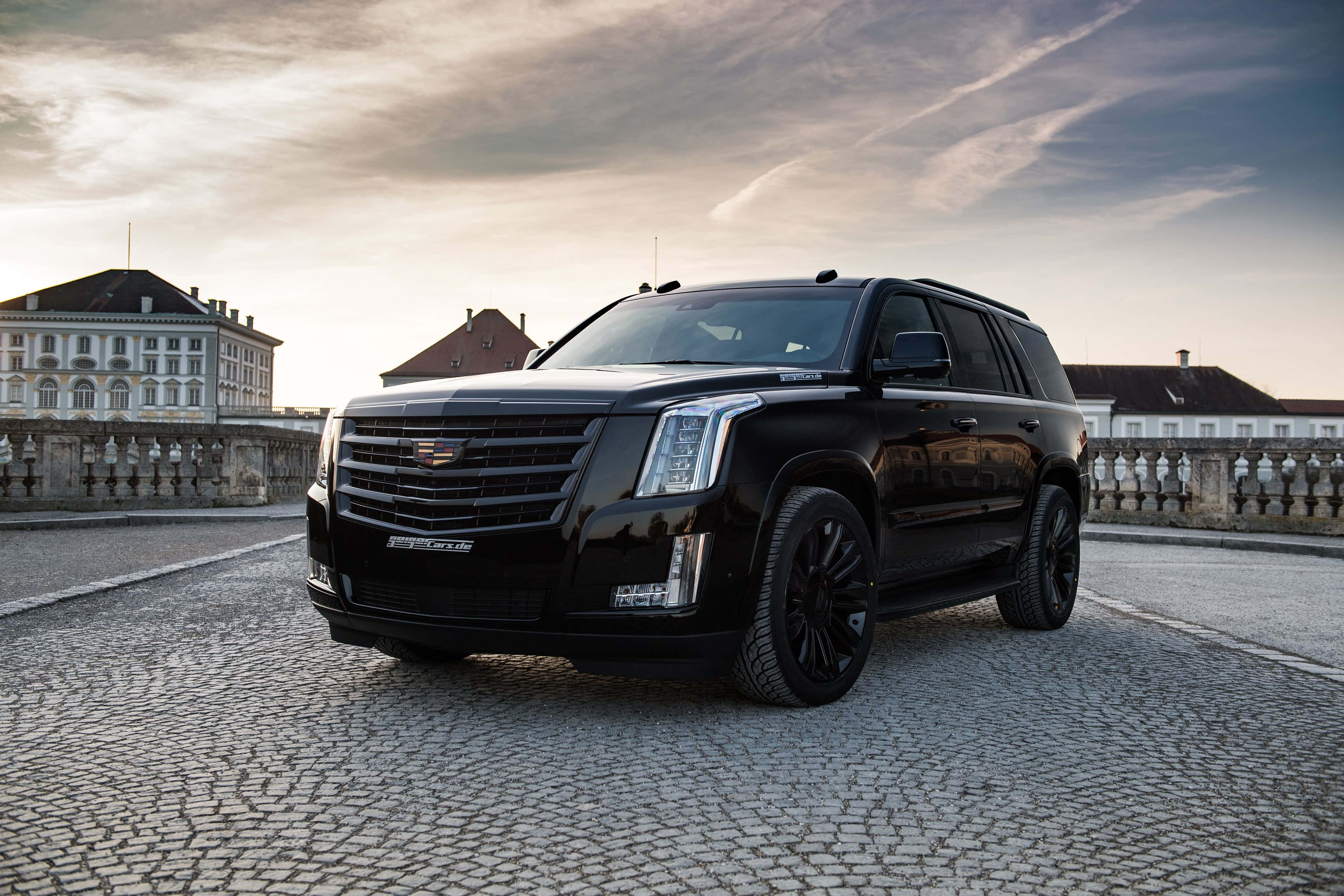 Cadillac Escalade 桌布 壁紙 — 一台時尚的黑色凱迪拉克 Escalade 停放在鵝卵石露台上，背景是歷史建築，天空呈現柔和的金色日落。