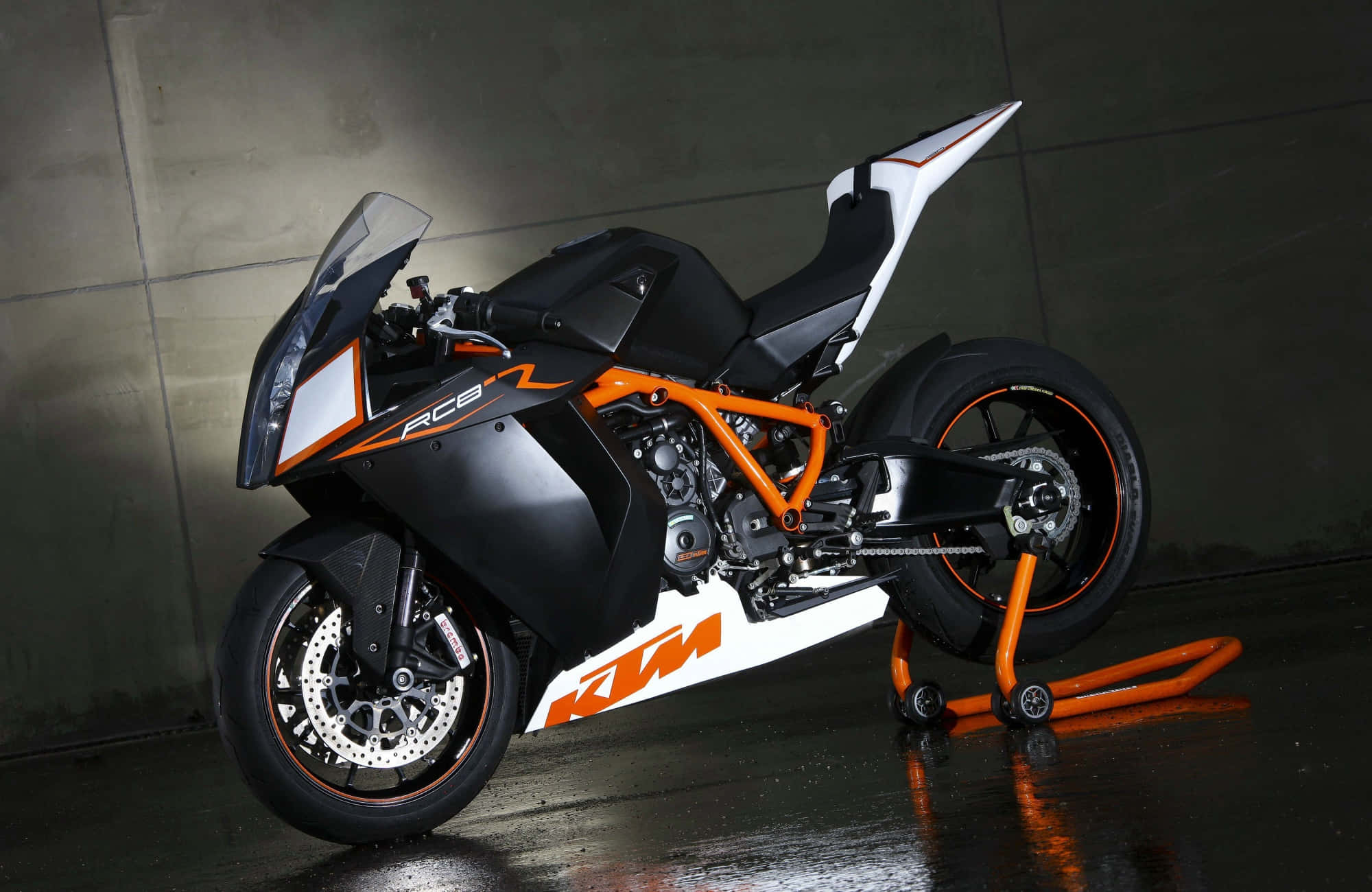 機車 桌布 壁紙 — 一台時尚的黑色和橙色KTM RC8摩托車停放在支架上，位於燈光昏暗的混凝土車庫中，地板潮濕反光。