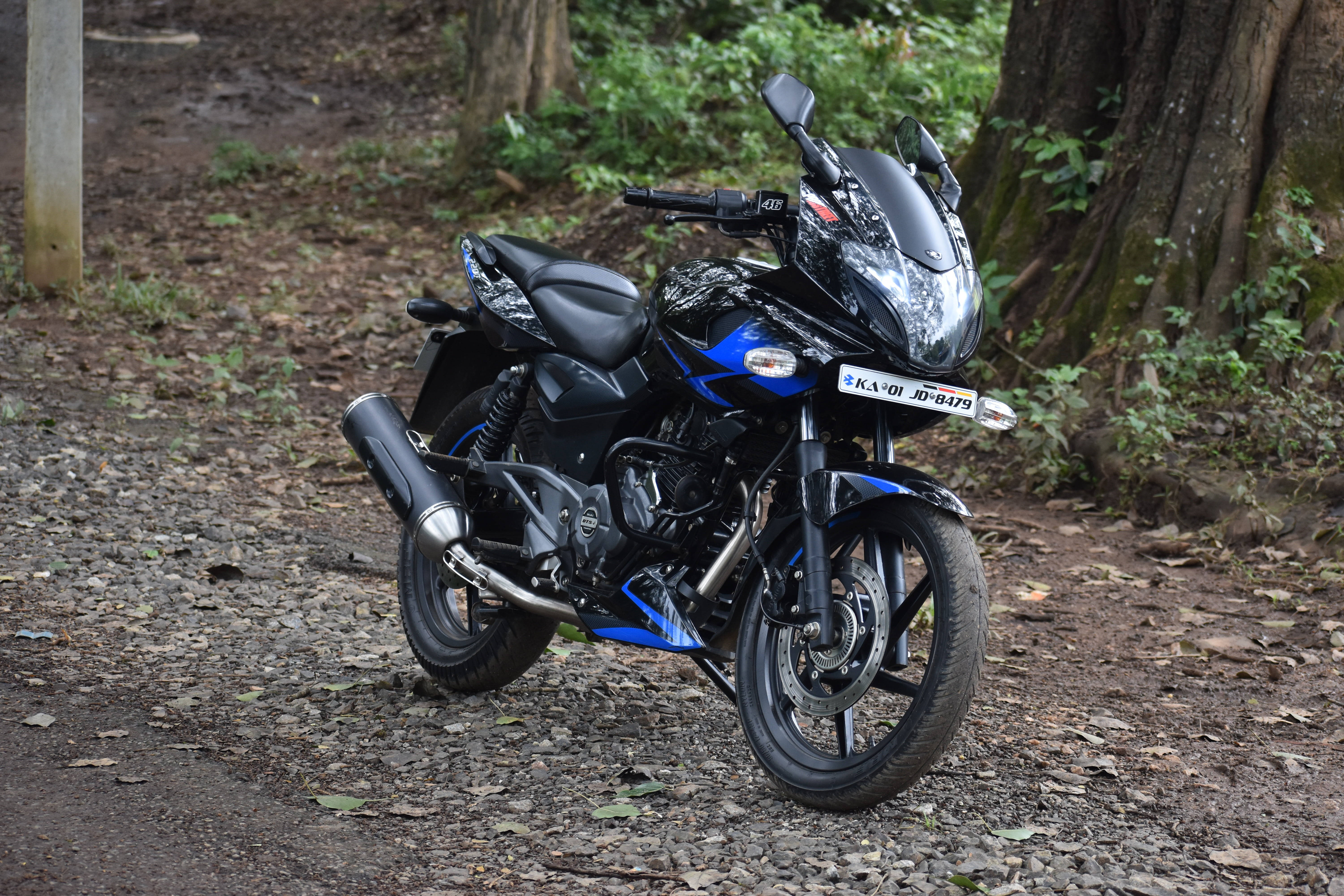 pulsar 220 HD 桌布 壁紙 — 一台時尚的黑色和藍色機車停放在森林碎石小徑上，周圍環繞著樹木和綠植。