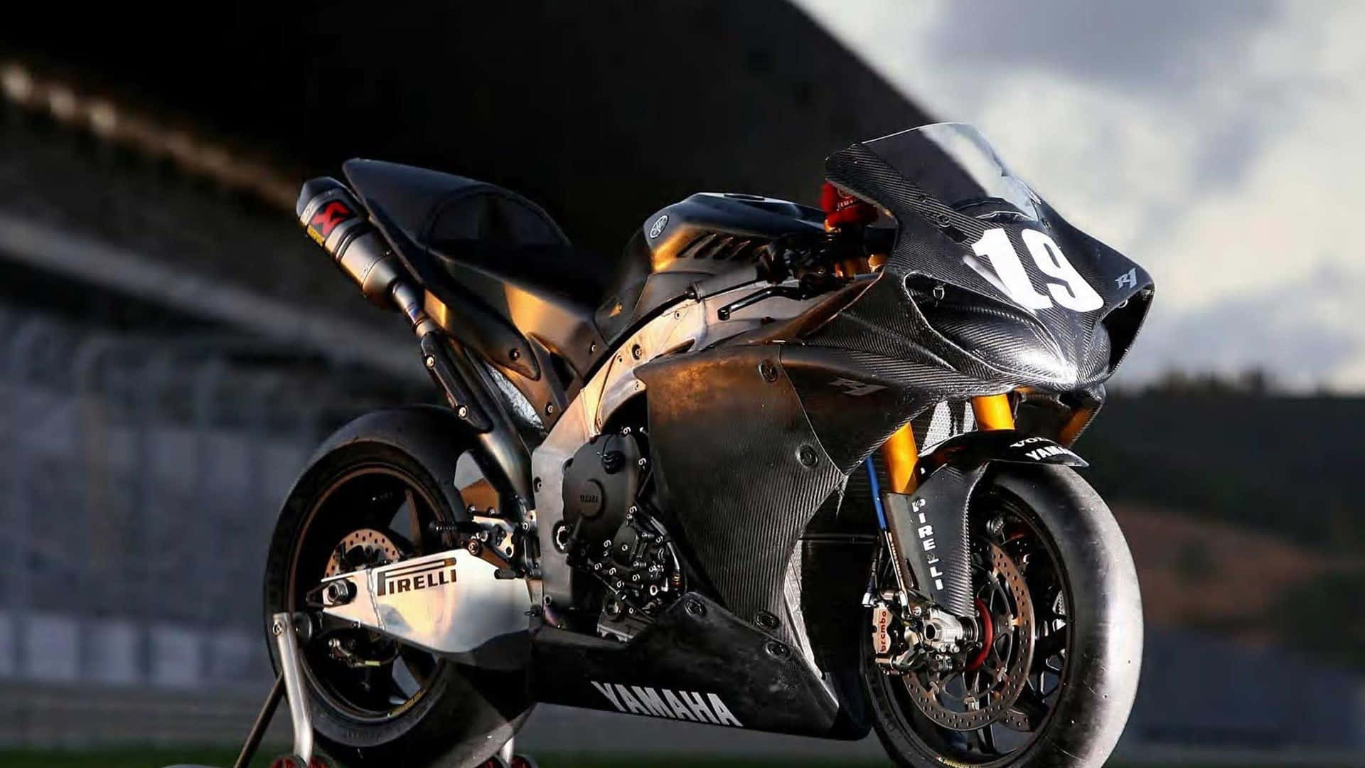 Superbike 桌布 壁紙 — 一台時尚的黑色和金色Yamaha賽車摩托車，號碼19，在戲劇性的天空燈光下停在賽道上。