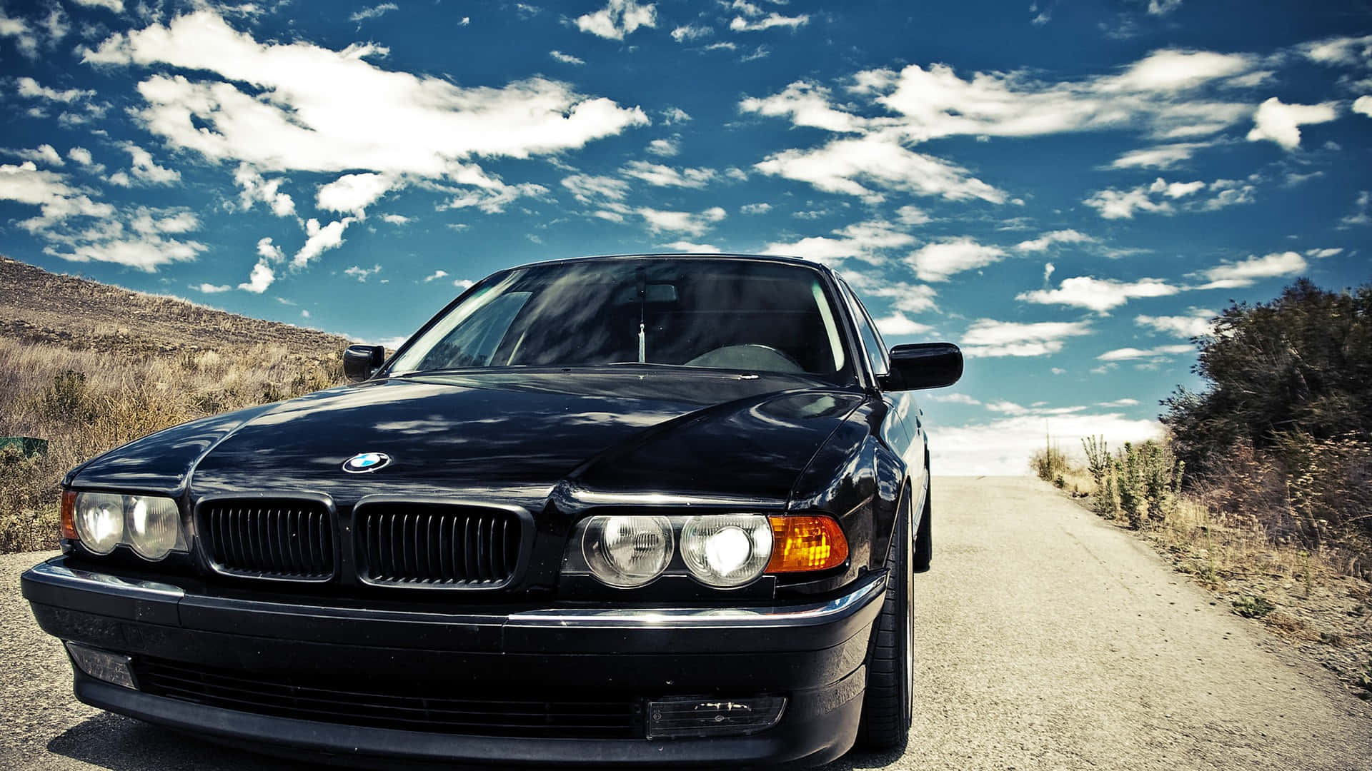 1920x1080 Bmw 桌布 壁紙 — 一台時尚的黑色寶馬轎車在陽光普照的沙漠公路上，襯托著充滿活力的藍天和蓬鬆的白雲。