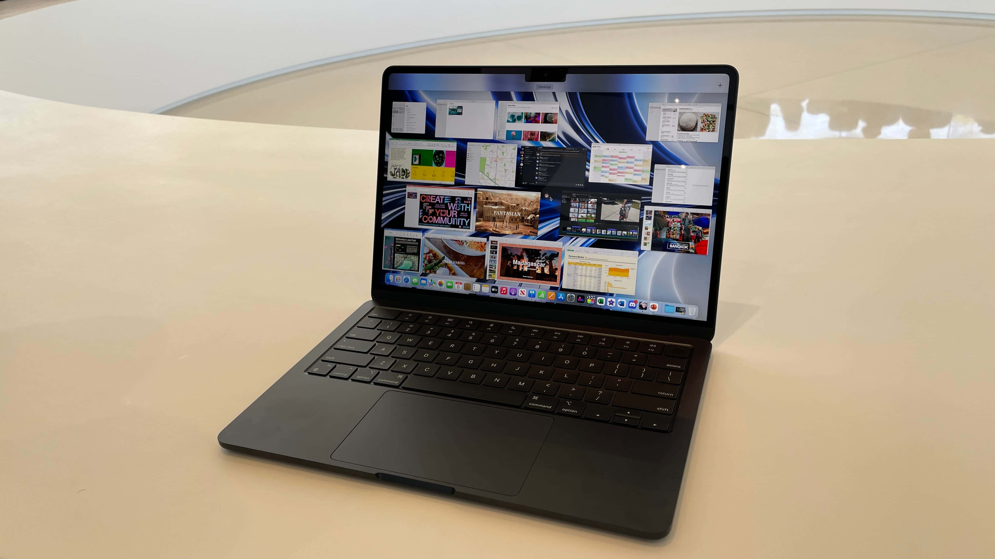 Black Macbook 桌布 壁紙 — 一台時尚的黑色筆記型電腦打開在淺米色表面上，顯示充滿活力的數位縮圖拼貼。