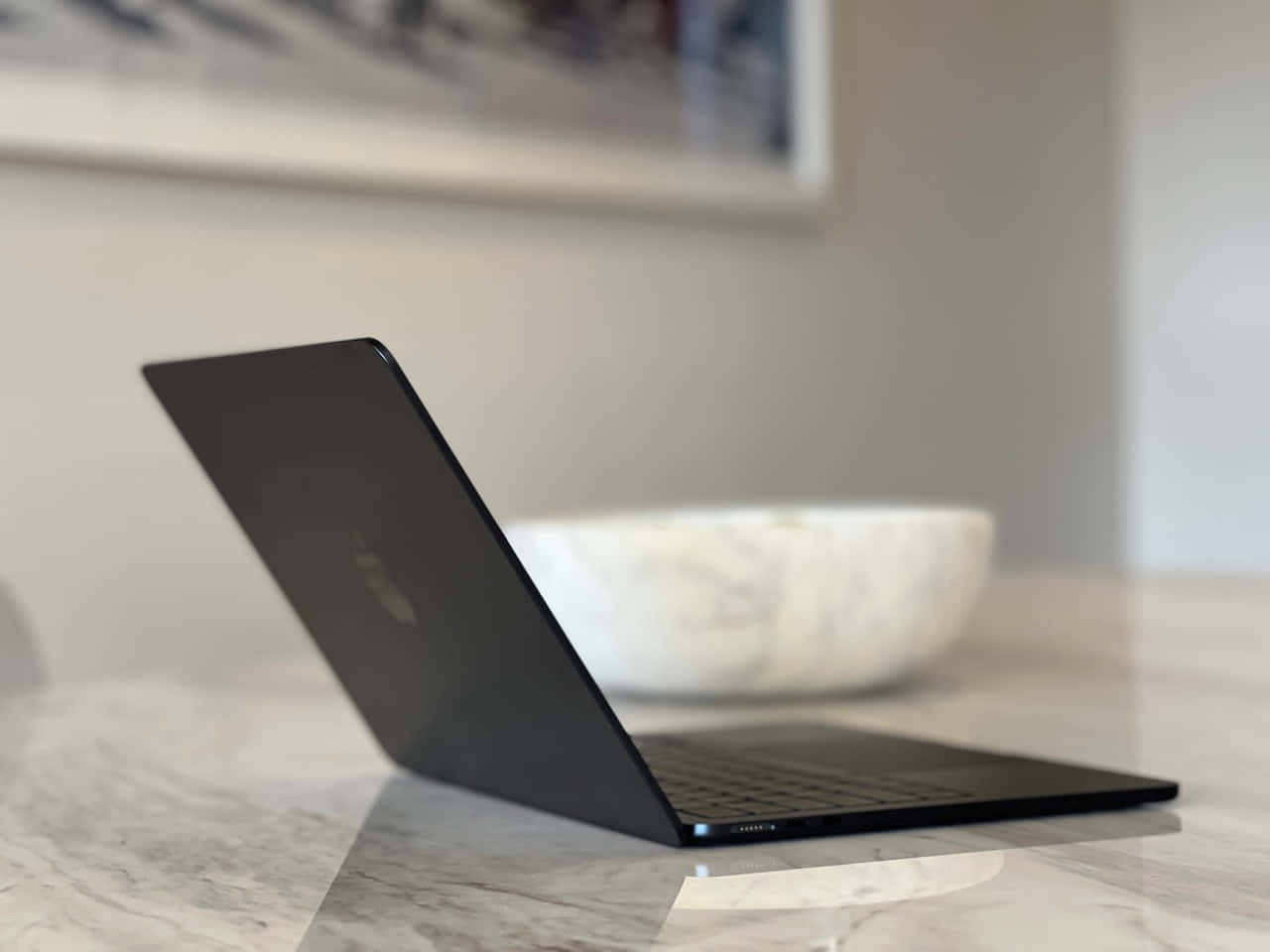 Black Macbook 桌布 壁紙 — 一台時尚的黑色筆記型電腦打開放在拋光大理石檯面上，背景柔和，有一個白色碗和裱框藝術品。