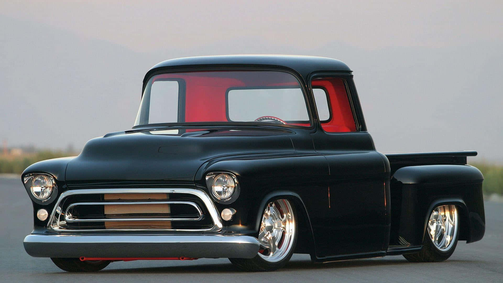 chevy 桌布 壁紙 — 一台時尚的黑色經典皮卡車，配備紅色內裝和鍍鉻裝飾，停放在開闊的道路上，背景是朦朧的山巒。